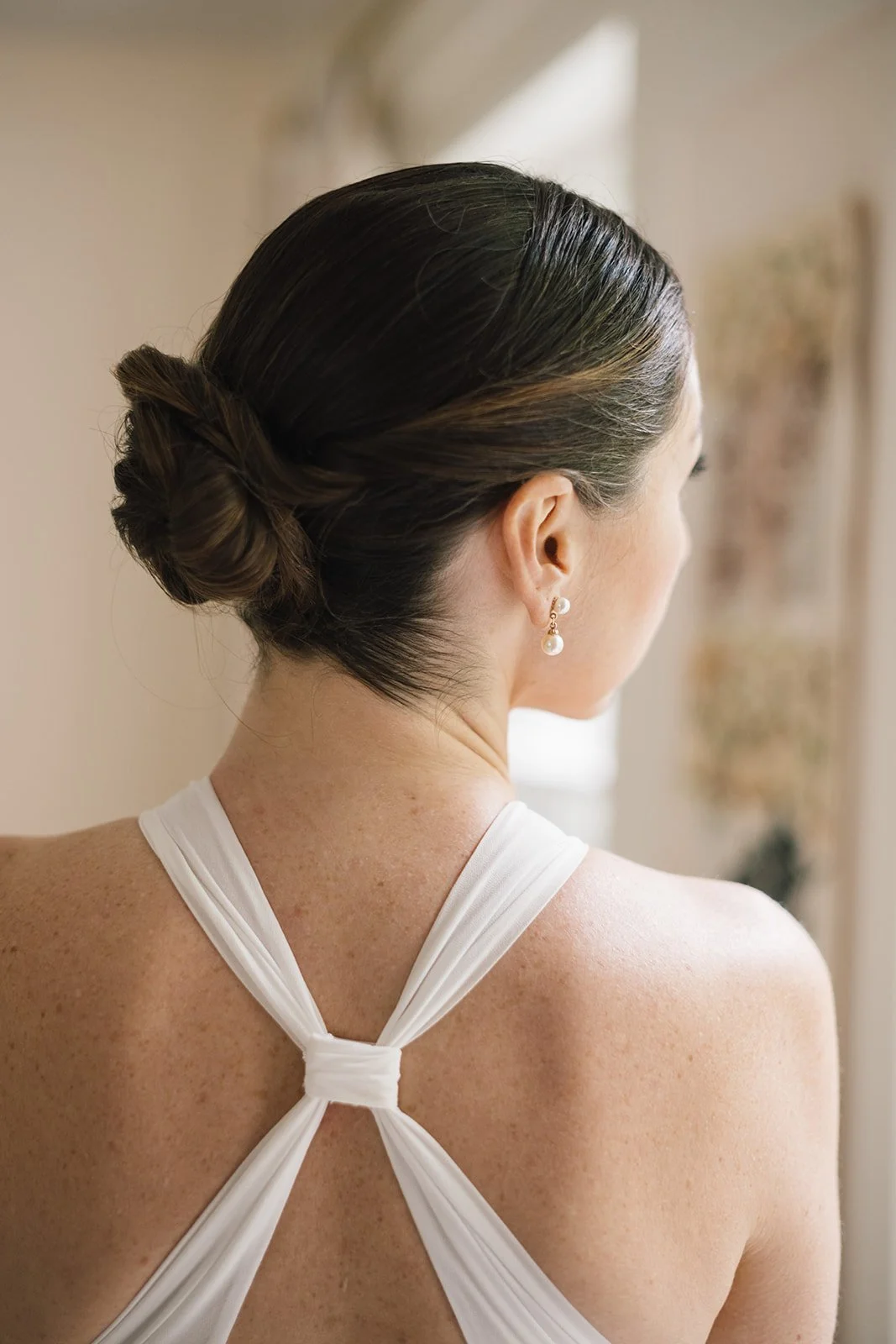 A woman with dark brown hair styled in an elegant updo, wearing pearl earrings and a white dress with a pleated back neckline, viewed from behind. Vermont Bridal Hairstyle. Wildflower Beauty Company. Burlington VT, Stowe, Manchester VT. Vermont Weddi