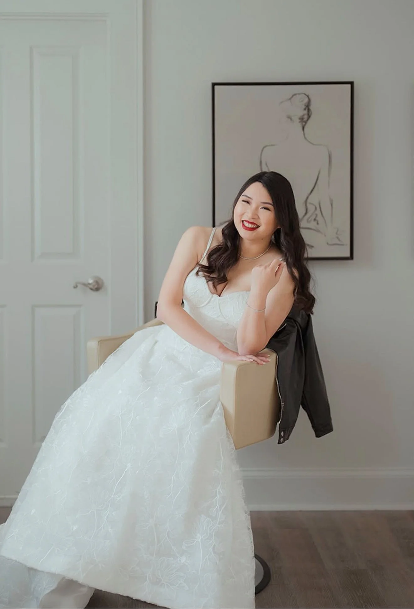 Beautiful Bride in a white wedding dress sitting on a chair, smiling and winking, with a black jacket draped on the chair's back, in a room with a white door and a minimalist line drawing of a seated woman on the wall behind her.