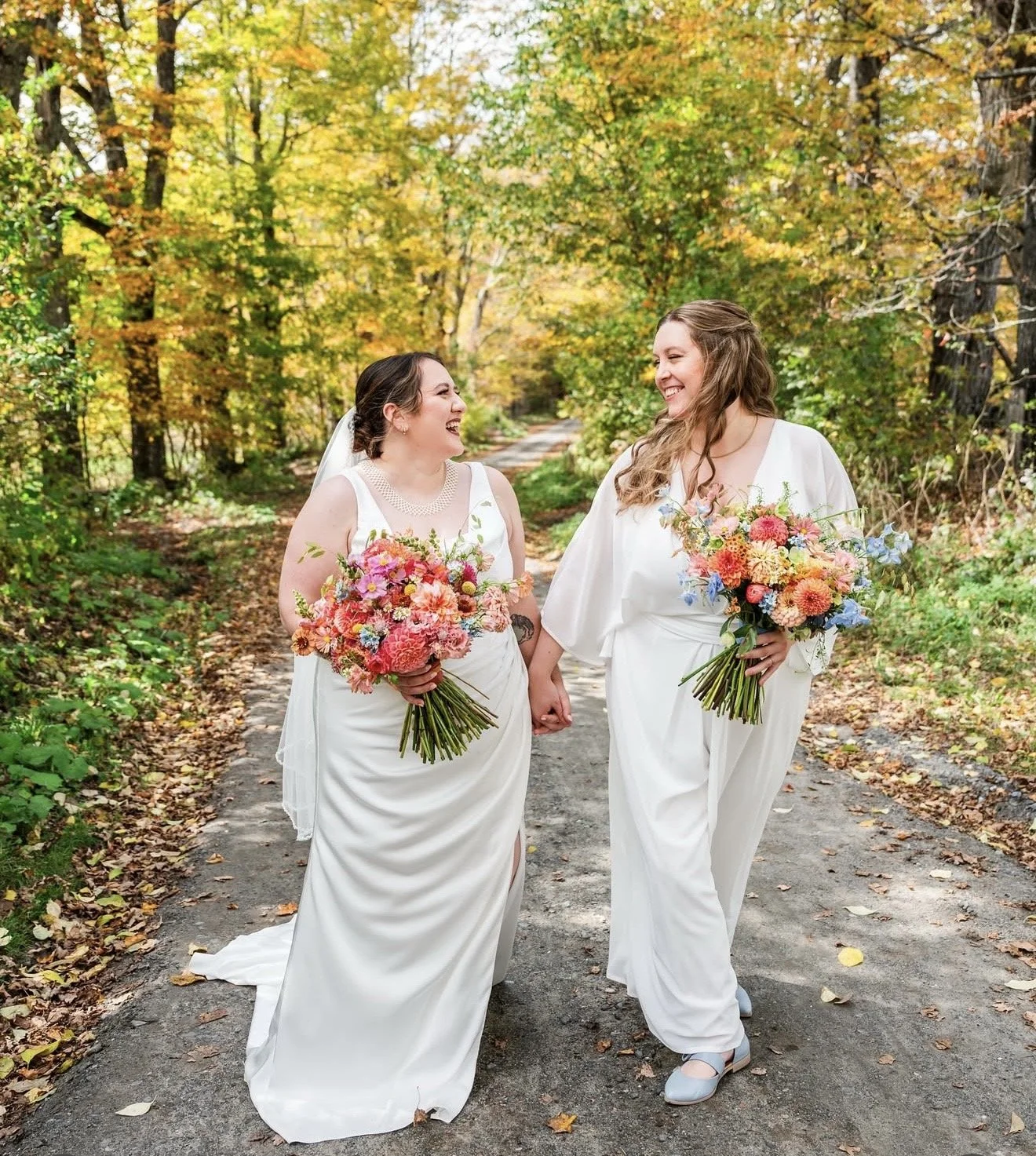 Two women in white dresses holding colorful bouquets, walking hand in hand along a dirt path surrounded by autumn foliage, smiling at each other. Vermont Bride. Vermont Bridal Hairstyle. Wildflower Beauty Company. Burlington VT, Stowe, Manchester VT