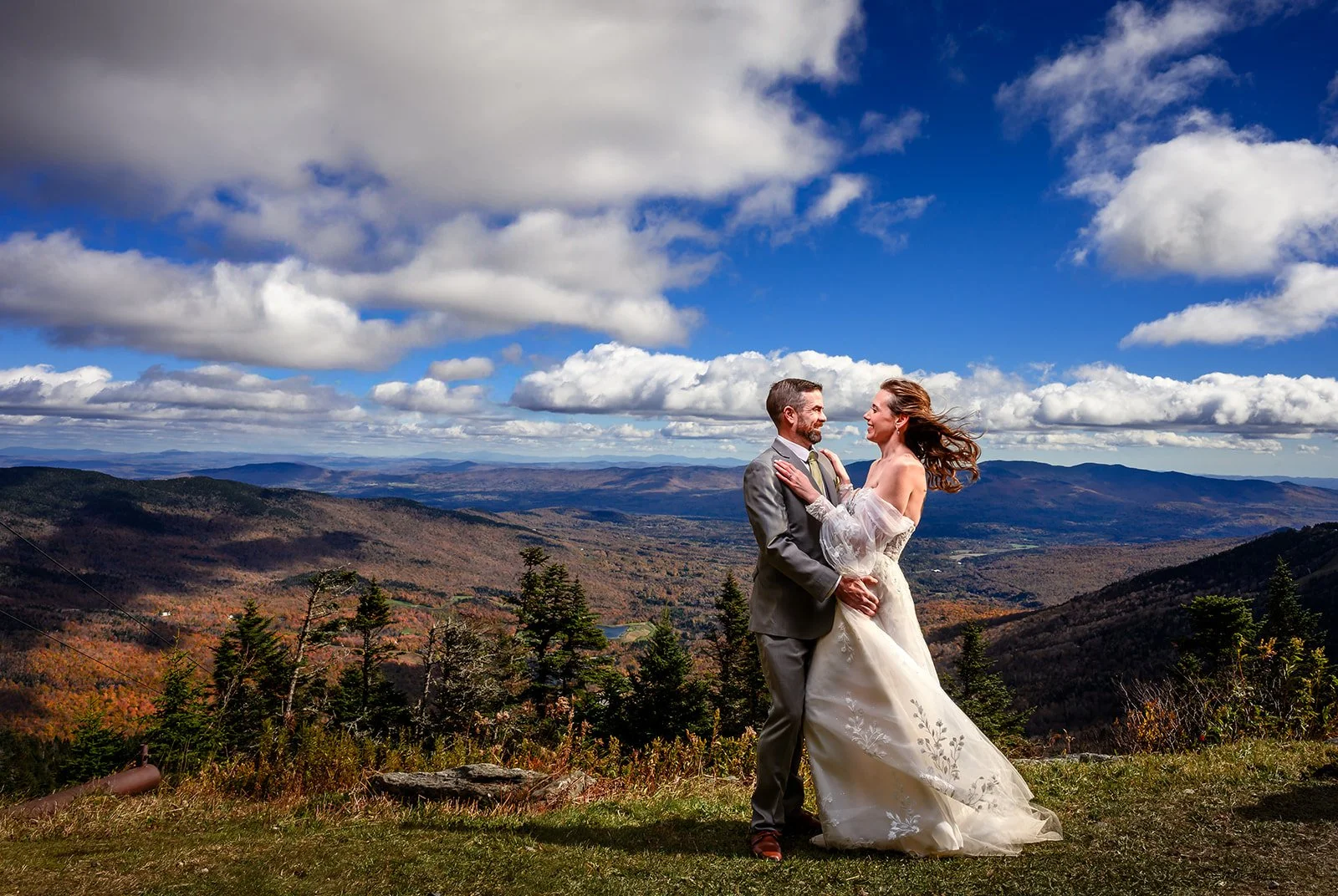 A couple in wedding attire standing on a grassy hill with a scenic mountain landscape and a cloudy sky in the background in Stowe Vermont at Spruce Peak