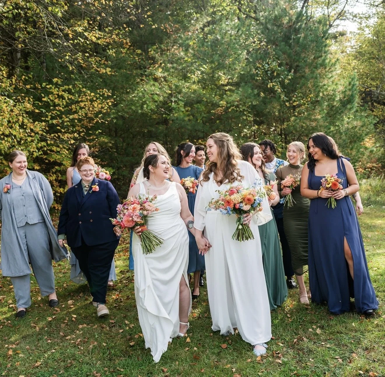 Group of women, including two brides, walking outdoors on a grassy area surrounded by autumn trees, holding flowers, and smiling. Vermont Bridal Hairstyle. Wildflower Beauty Company. Burlington VT, Stowe, Manchester VT. Vermont Wedding, Vermont Bride