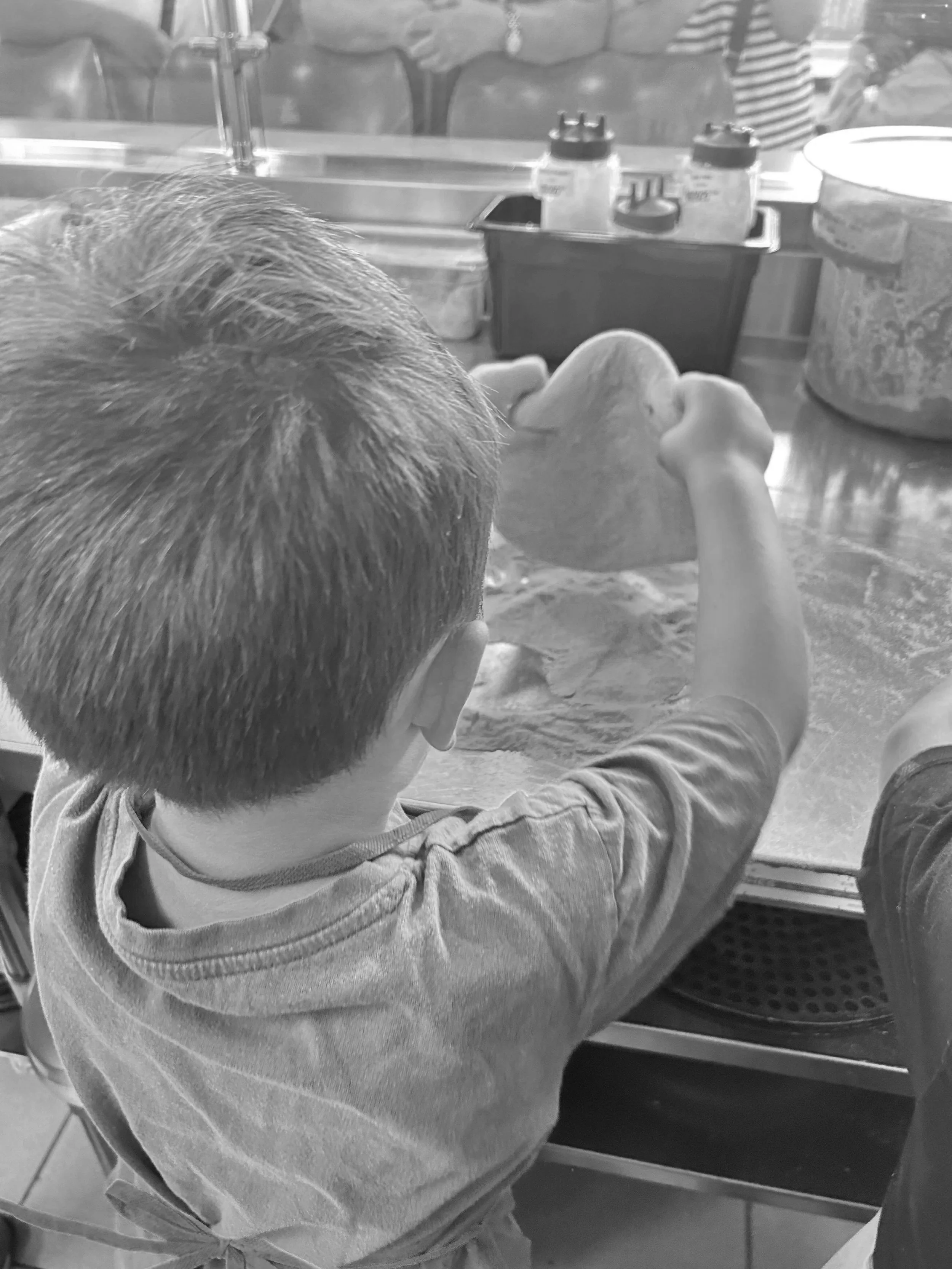 A young boy wearing a striped shirt is sitting at a counter, focused on making a pizza with a dough ball in his hands, surrounded by a pizza-making station with various toppings and tools. Location 1555 Talbot Rd, Lasalle, Ontario.