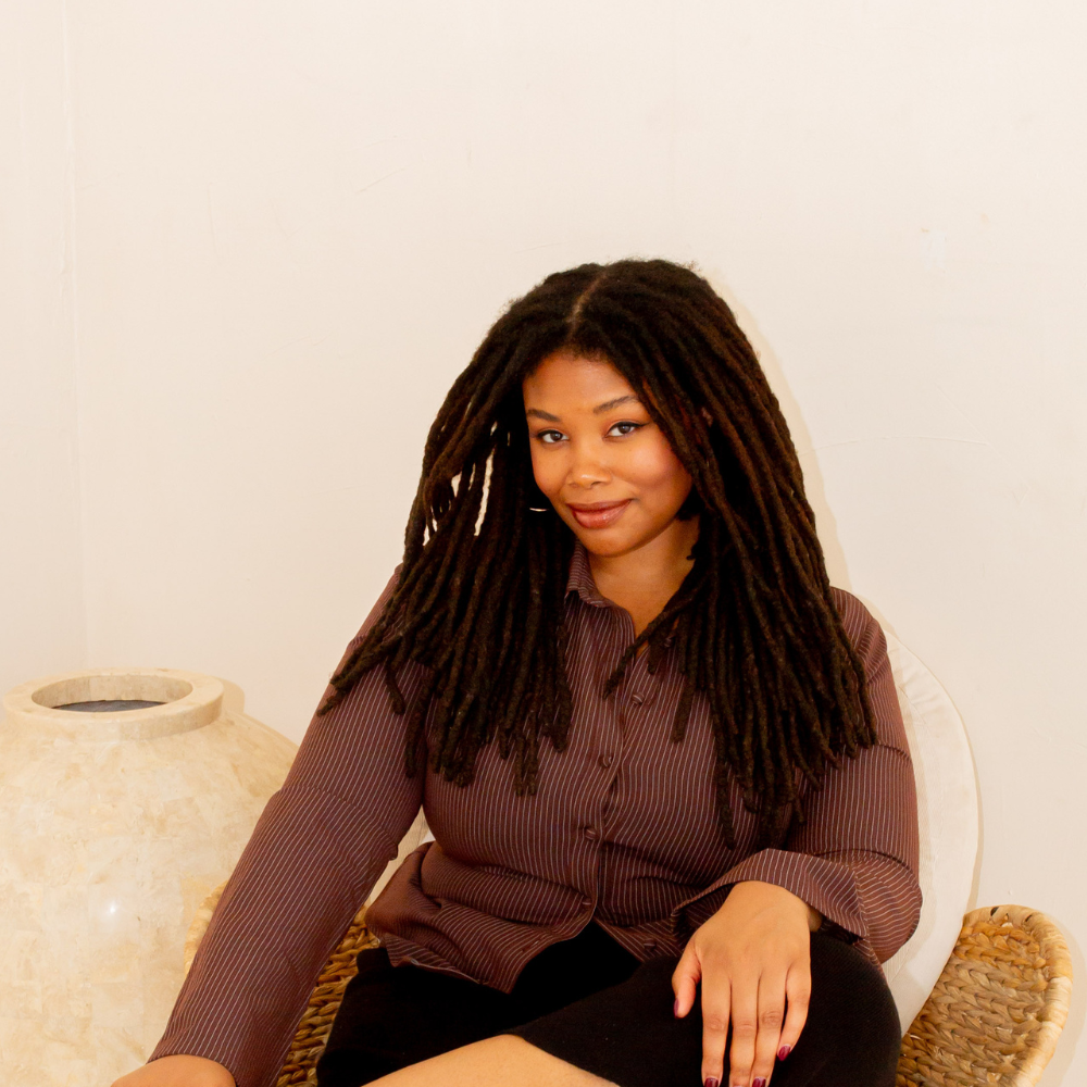 A woman with long dreadlocks, wearing a brown pinstriped blouse, sitting on a wicker chair with a cream cushion, in front of a neutral-colored wall.