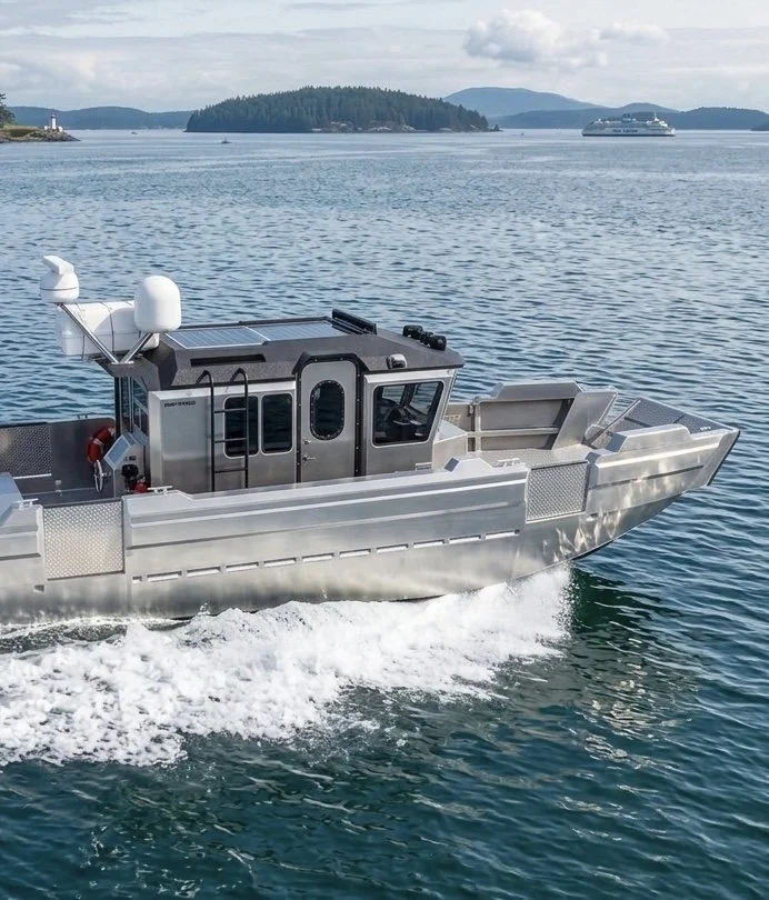 A metallic boat with a cabin is sailing on a body of water, with land and a cruise ship visible in the background.