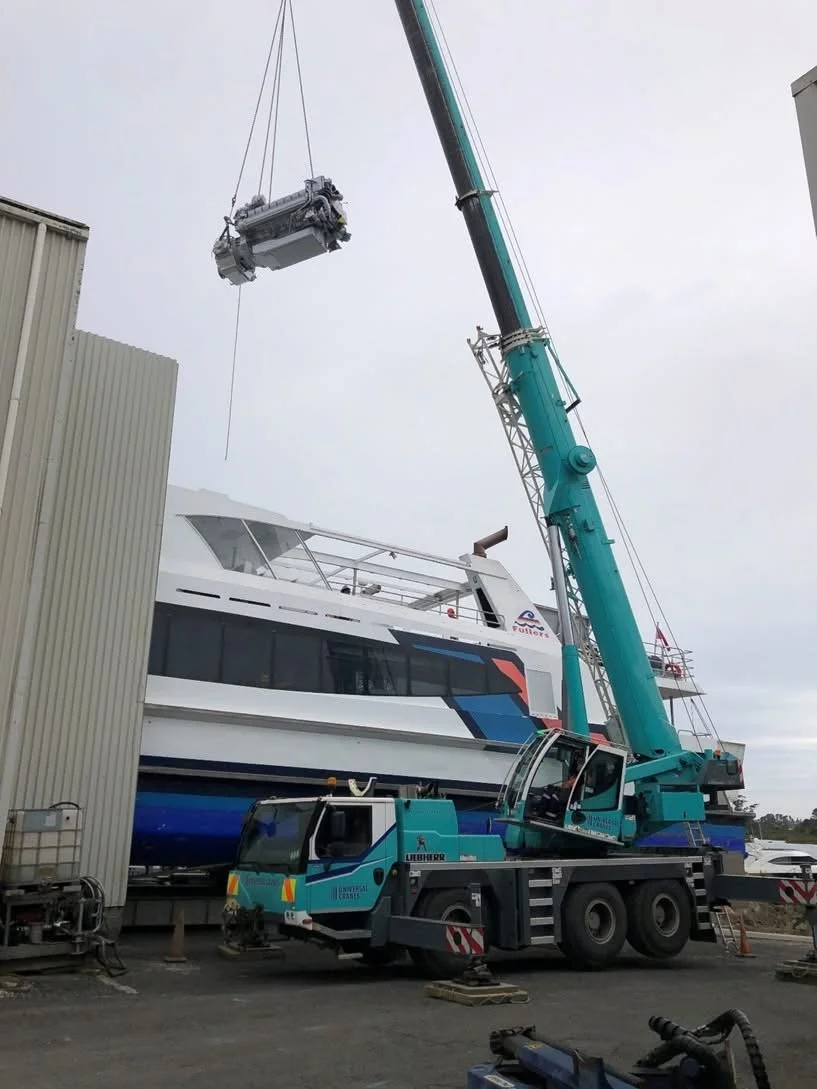 A large blue crane lifting a heavy engine or machinery part over a boat docked at a marina, with construction equipment visible around.