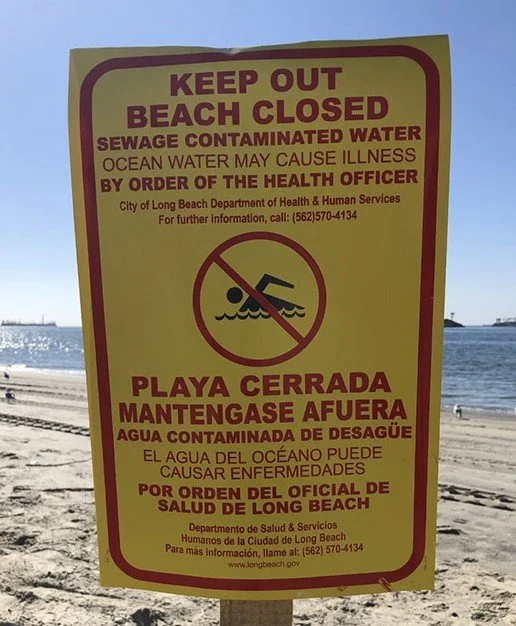 Sign on a beach warning about sewage contaminated water, restricting access and indicating health risks in English and Spanish.