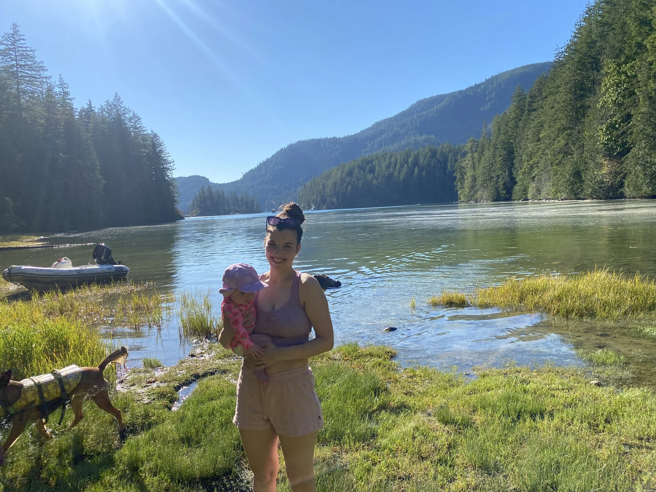A woman holding a child by a lake with a forested shoreline and mountains in the background, a boat on the water, and a dog on a leash near the grass on a sunny day.