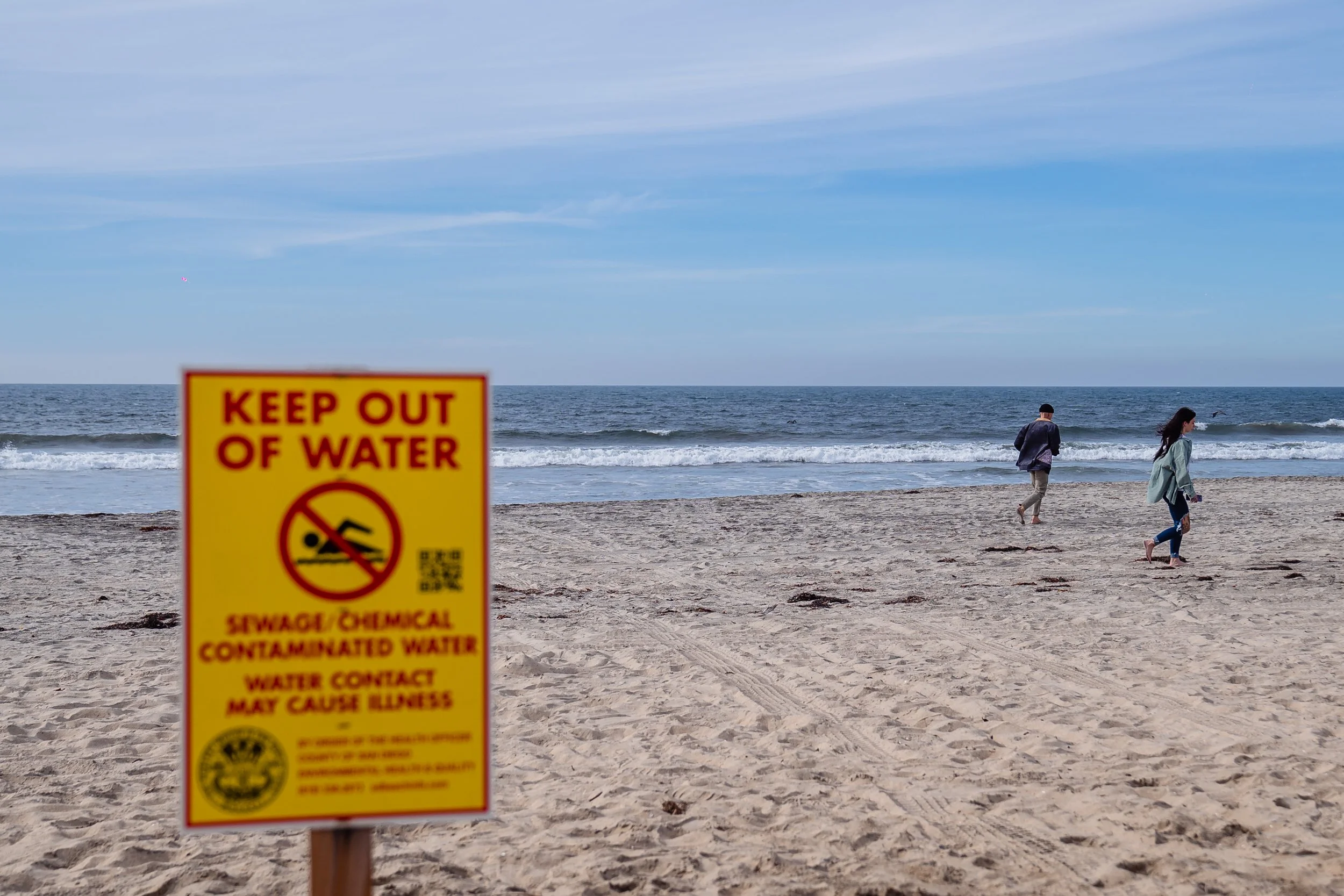A beach with two people walking along the shoreline, and a yellow warning sign in the foreground indicating to keep out of water due to sewage and chemical contamination.