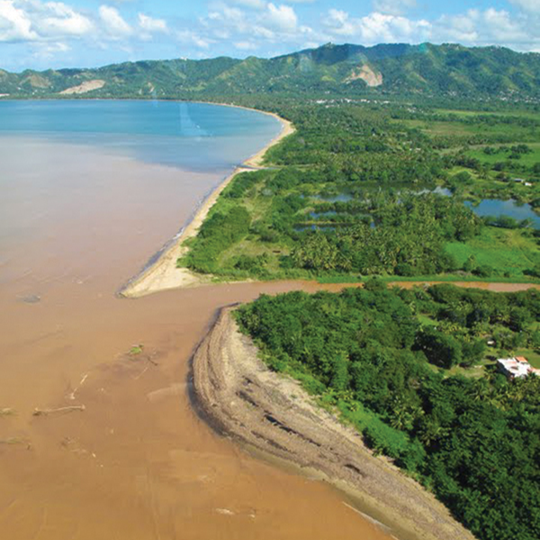 Aerial view of a coastal area with a sandy beach, green vegetation, and water bodies, with mountains in the background.