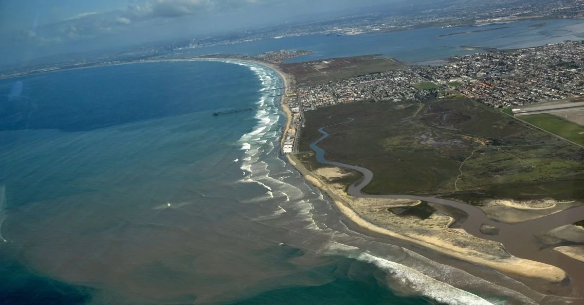 Aerial view of a coastal city with a beach, ocean waves, a river, and urban landscape with buildings and roads.