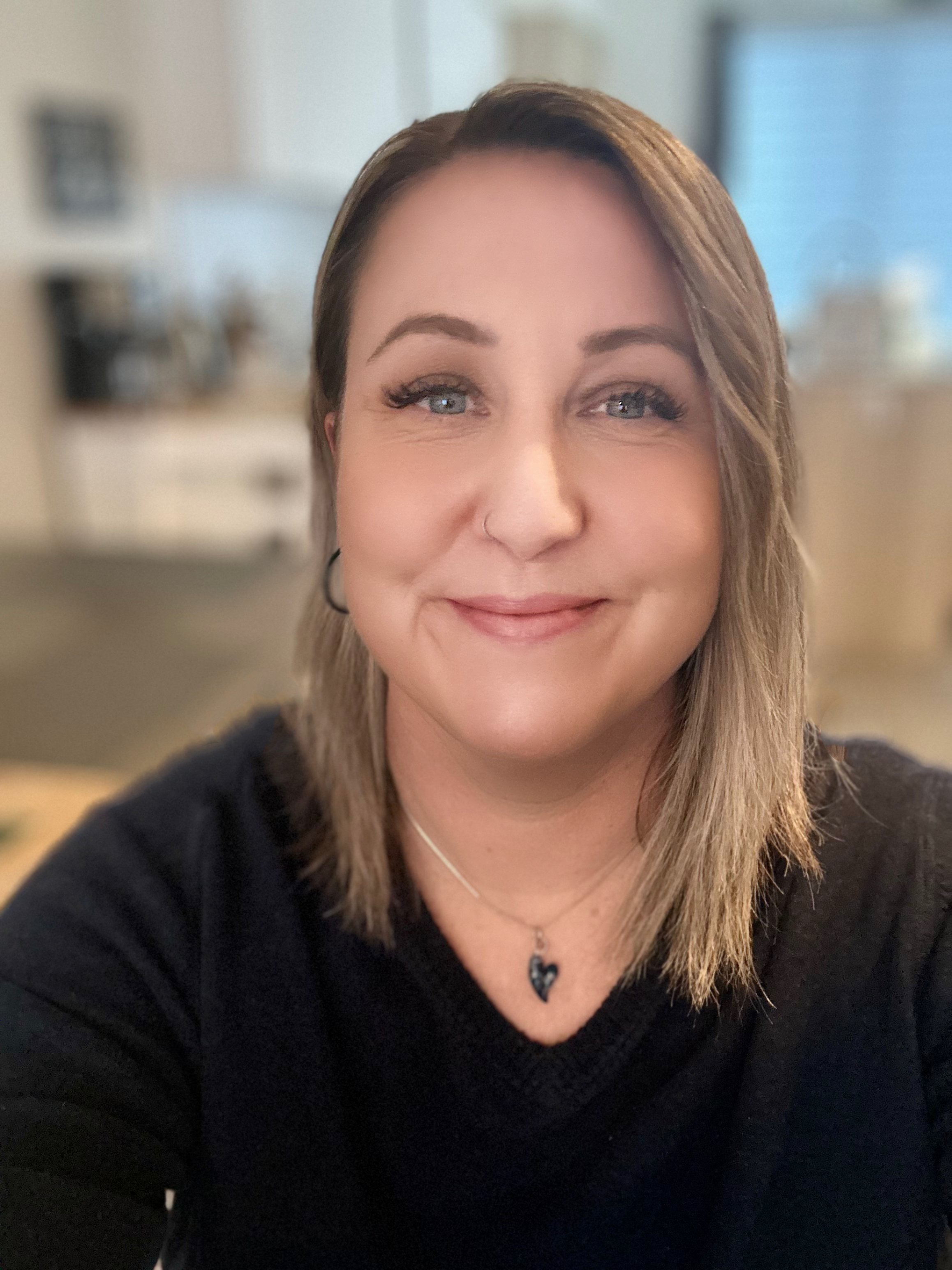 A woman with shoulder-length light brown hair, blue eyes, and fair skin, smiling at the camera, wearing a black top, a heart-shaped necklace, and hoop earrings, with a blurred kitchen background.