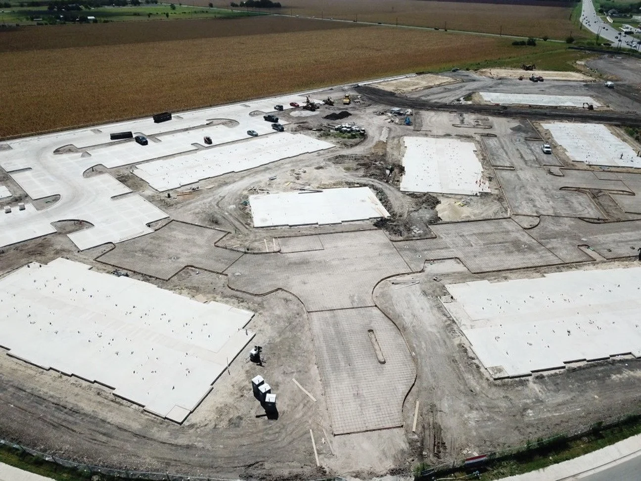 Aerial view of a construction site with partially built structures, parking area, construction vehicles, and surrounding farmland.