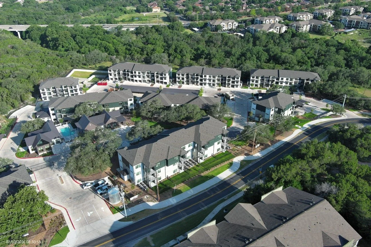Aerial view of a residential apartment complex with multiple buildings, swimming pools, parking lots, surrounded by green trees and a curving road.