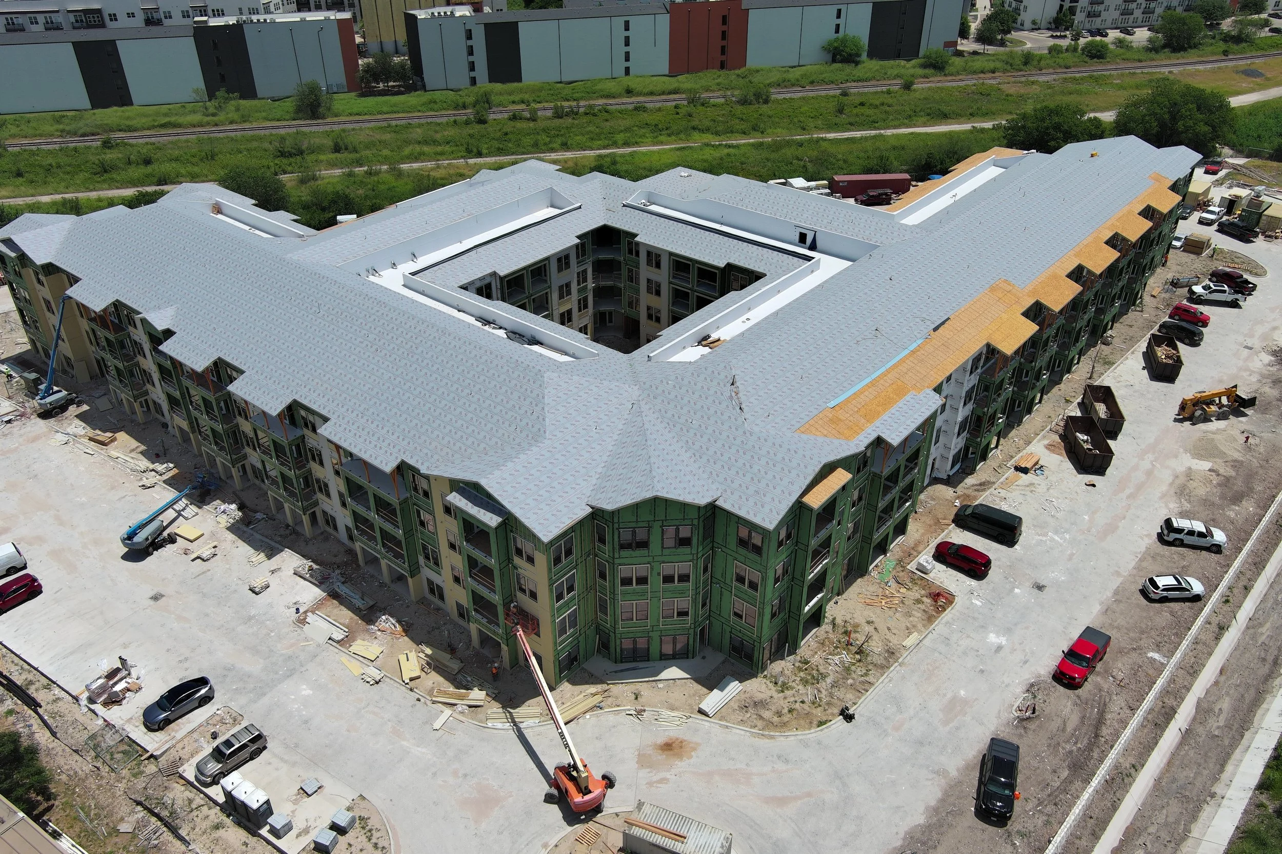 An aerial view of a large multi-story apartment complex under construction, with scaffolding and construction equipment visible, surrounded by parked cars and green space.