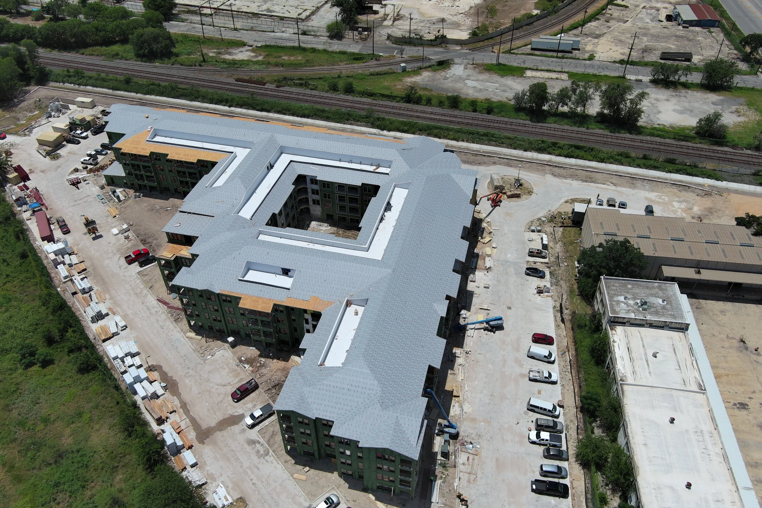 Aerial view of a construction site with a multi-story building under development, surrounding dirt areas, parked vehicles, and nearby railway tracks.