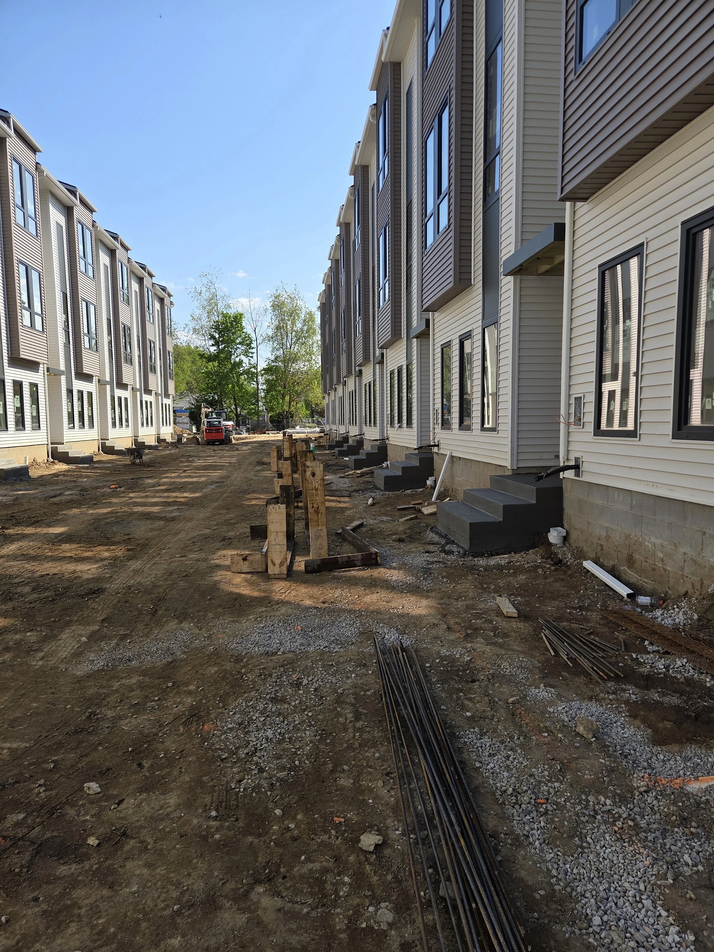 View of new construction apartment buildings with steps leading to entrances, construction materials and equipment on dirt ground, and trees in the background under a clear blue sky.