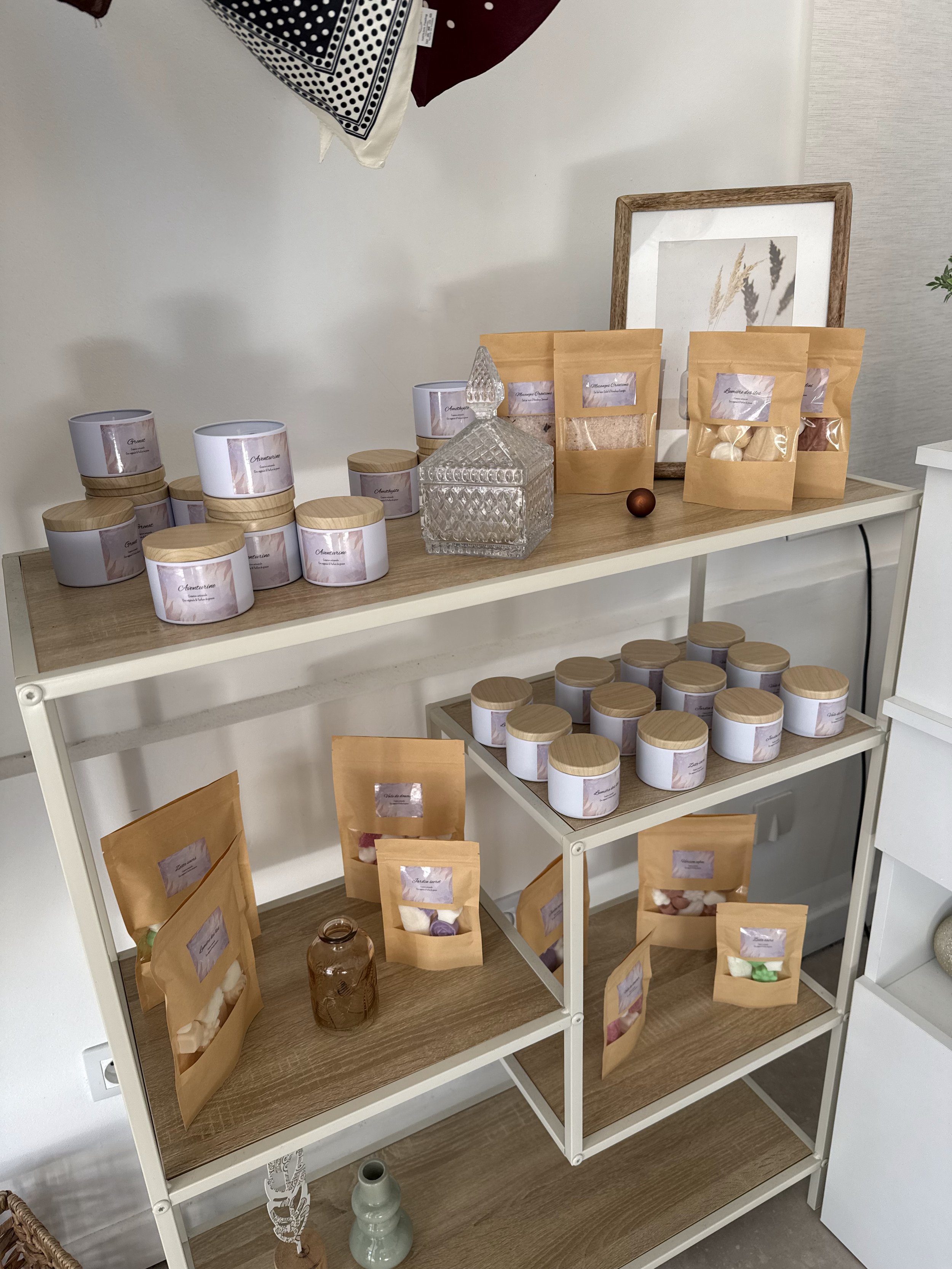 Une étagère en métal blanc contenant plusieurs pots en verre avec couvercle en bois, sacs en papier kraft transparent avec des étiquettes, un bocal en verre et une décoration en bois avec un plastron en céramique. Le tout dans un intérieur moderne avec un cadre en bois avec une impression de millet en arrière-plan.