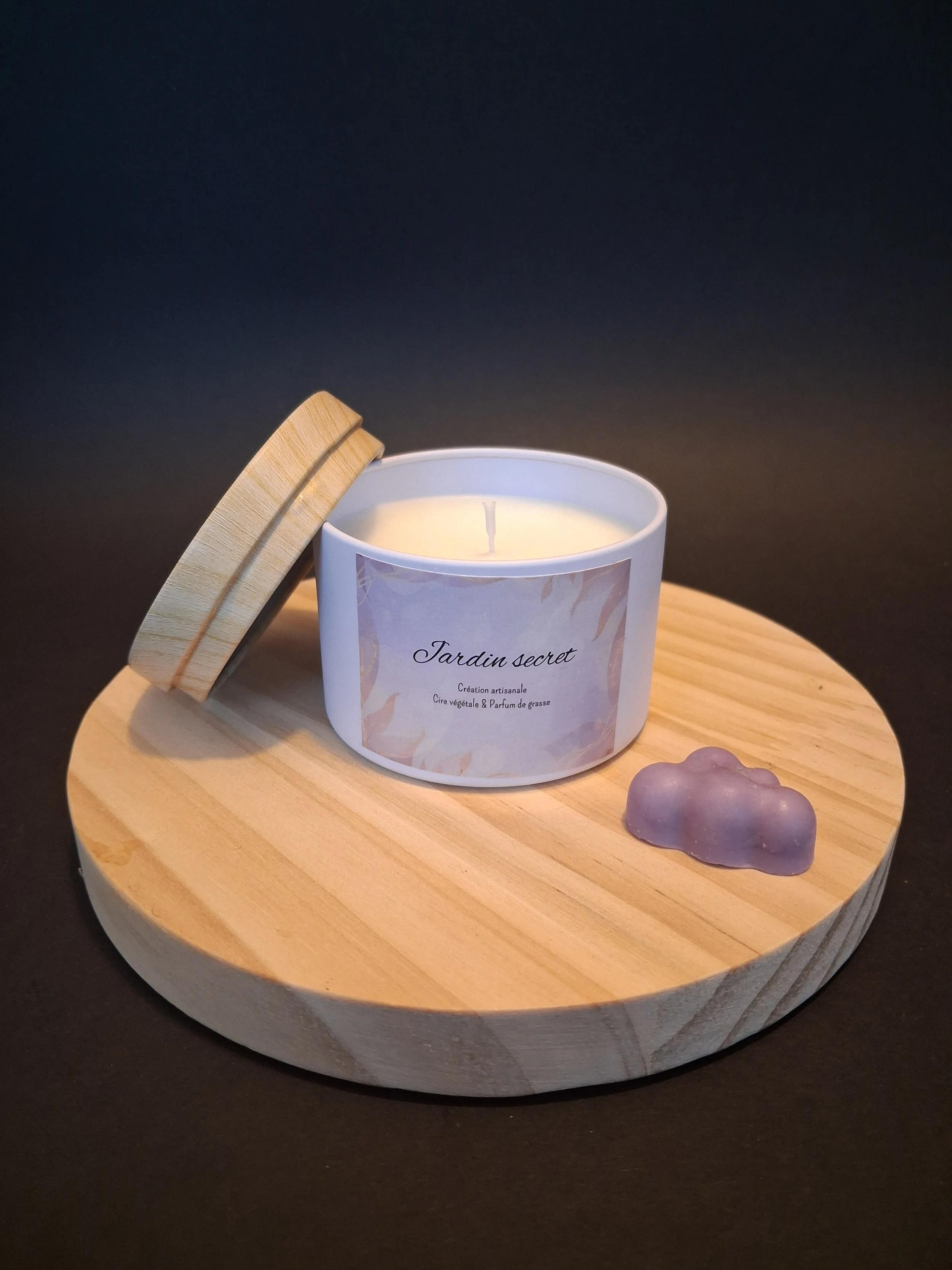Bougie blanche dans un récipient en céramique avec étiquette 'Jardin secret', accompagnée d'une petite figurine en forme de nuage violet clair, posées sur un plateau en bois rond, fond noir.
