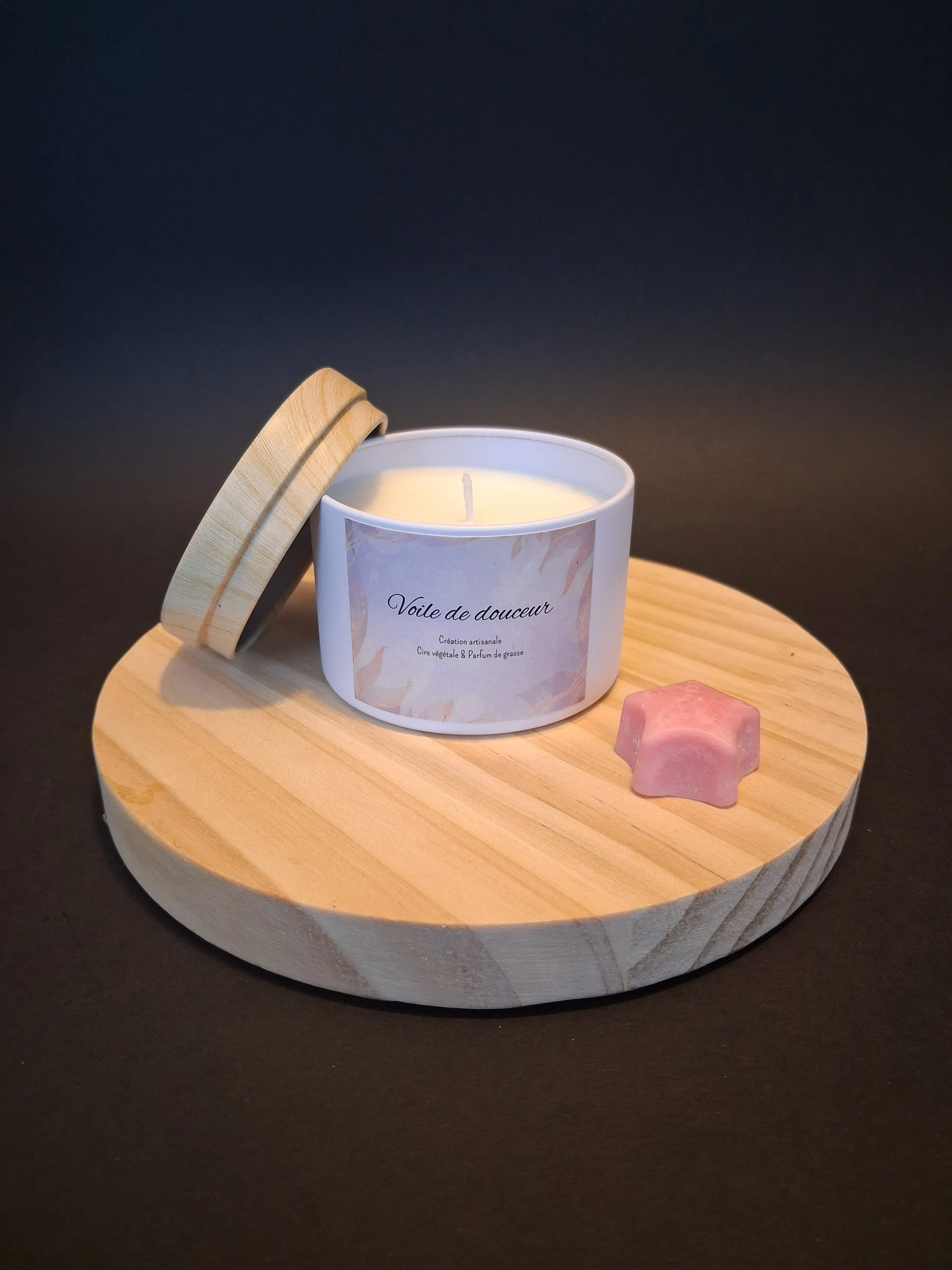 Bougie blanche dans un contenant avec une étiquette 'Voile de douceur', une boîte en bois avec un couvercle posé à côté, et une petite étoile en forme de savon rose sur un plateau en bois