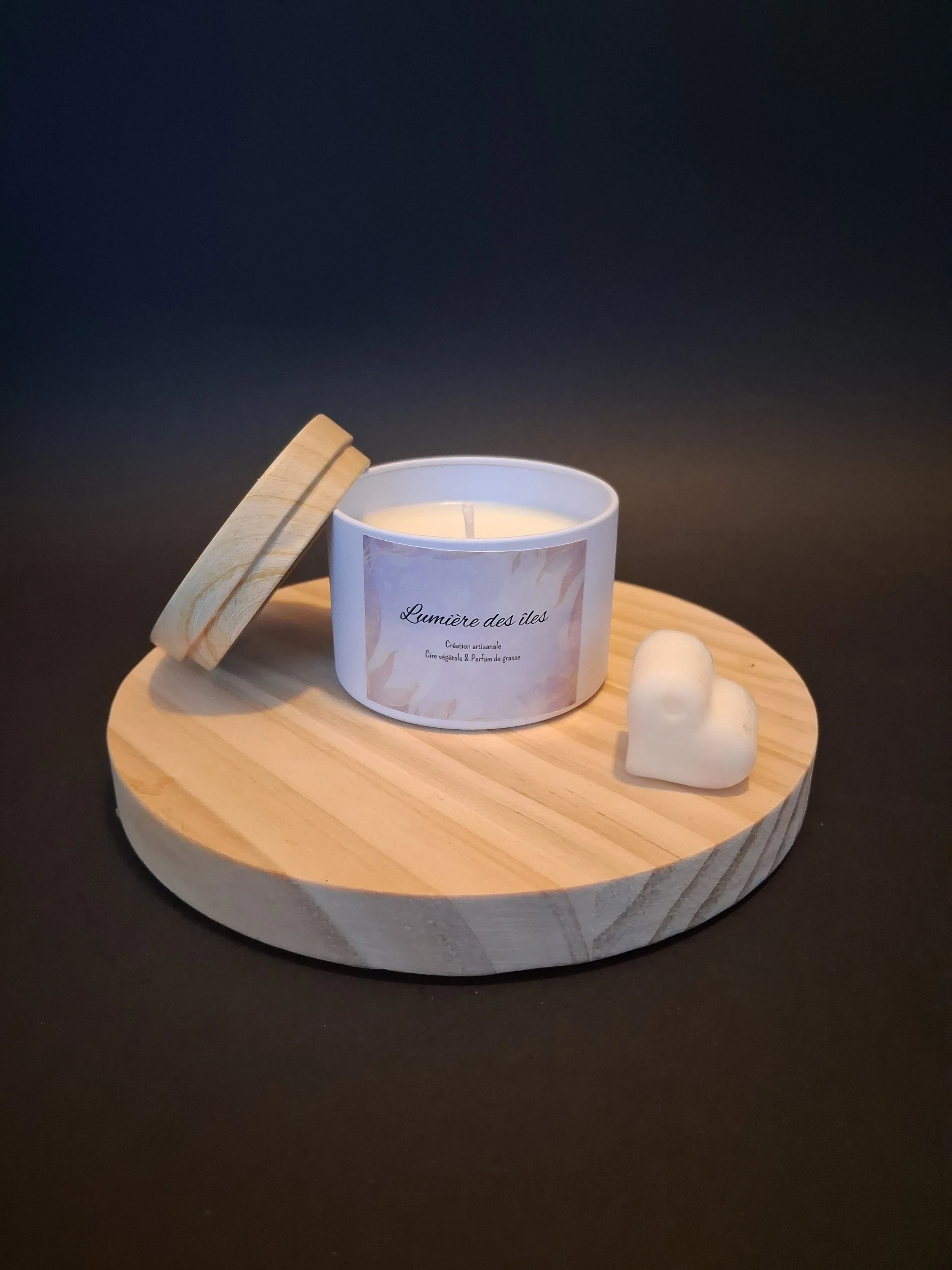 Bougie allumée dans un récipient blanc avec une étiquette, accompagnée d'une petite bougie en forme de cœur et d'un couvercle en bois, disposés sur une planche en bois sur fond sombre.