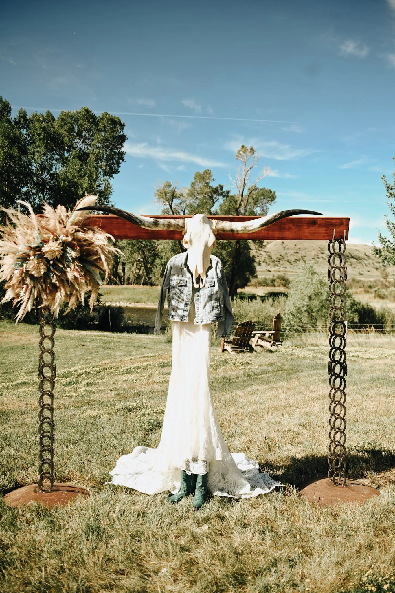 A rustic outdoor sculpture of a long dress and denim jacket on a mannequin with a cow skull and long horns mounted on a wooden beam, decorated with a bouquet of pampas grass and flowers, in a grassy field with trees and benches in the background under a partly cloudy sky.
