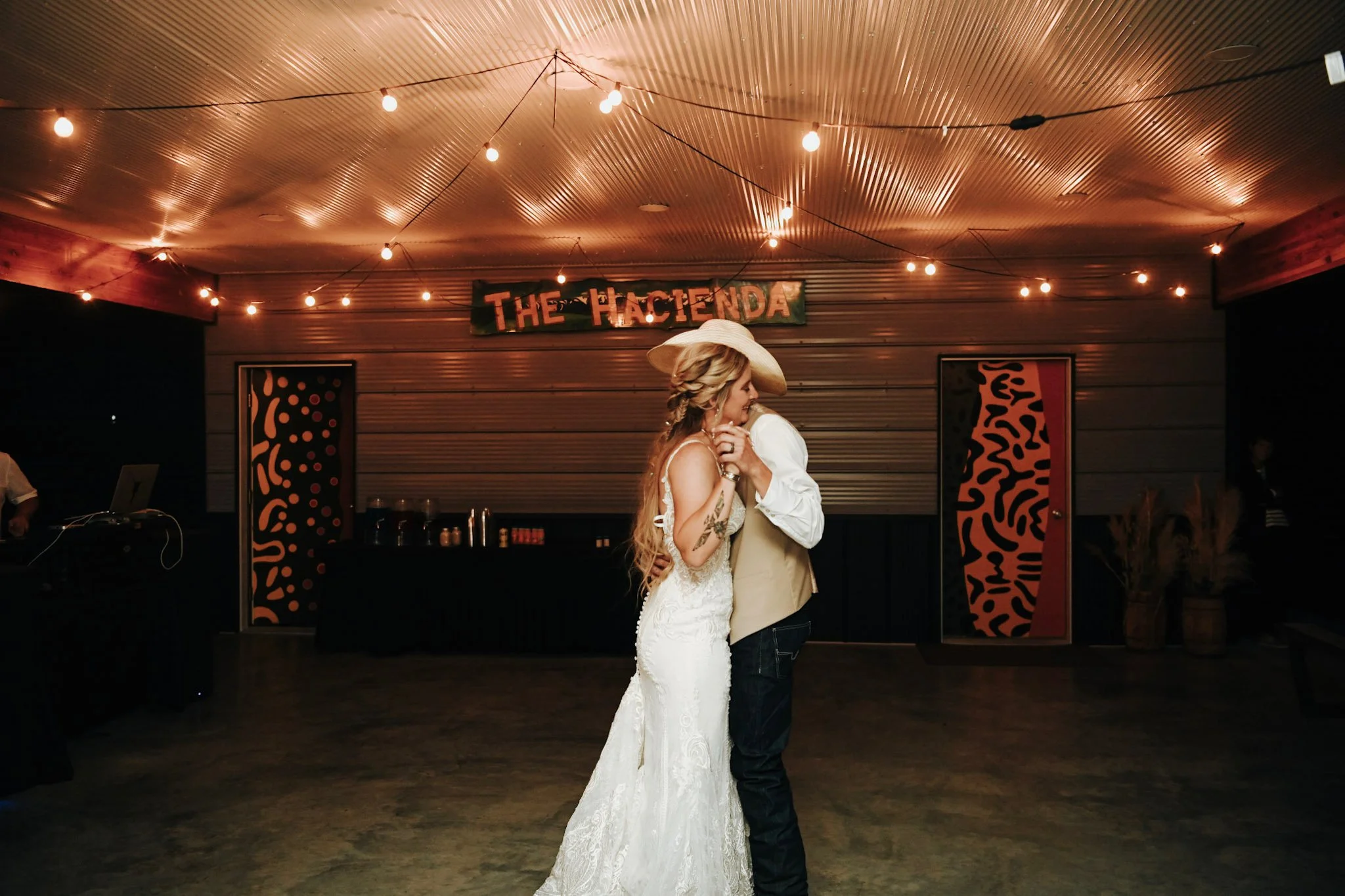 A couple dancing at a wedding reception, with the bride in a white lace dress and the groom in a beige vest and hat, under string lights and a sign that reads 'The Hacienda'.