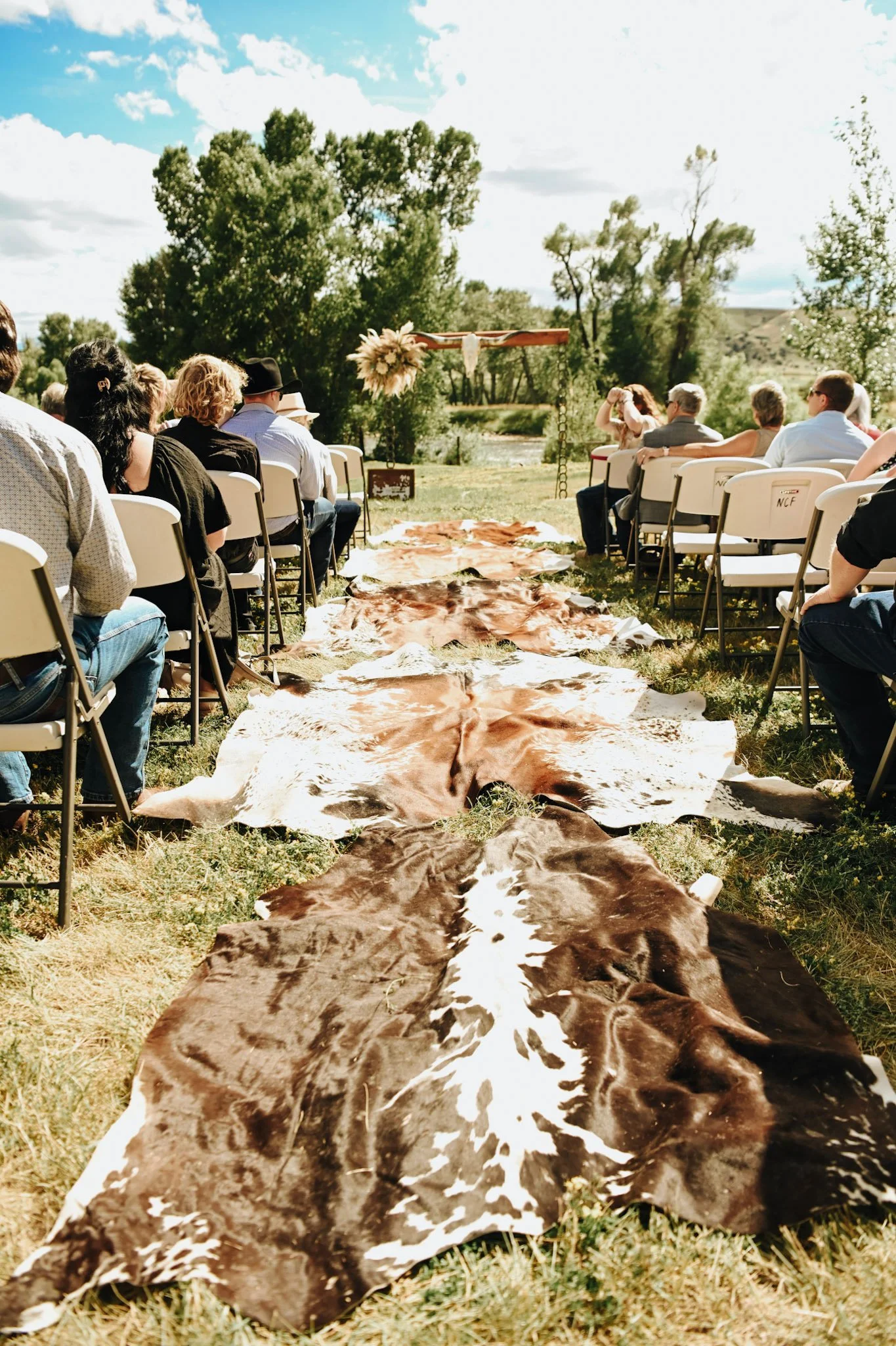 A group of people sitting on white chairs outdoors during a wedding ceremony, with cowhide rugs laid out on the grass in a line leading to an altar decorated with flowers and a wooden cross, surrounded by trees under a partly cloudy sky.