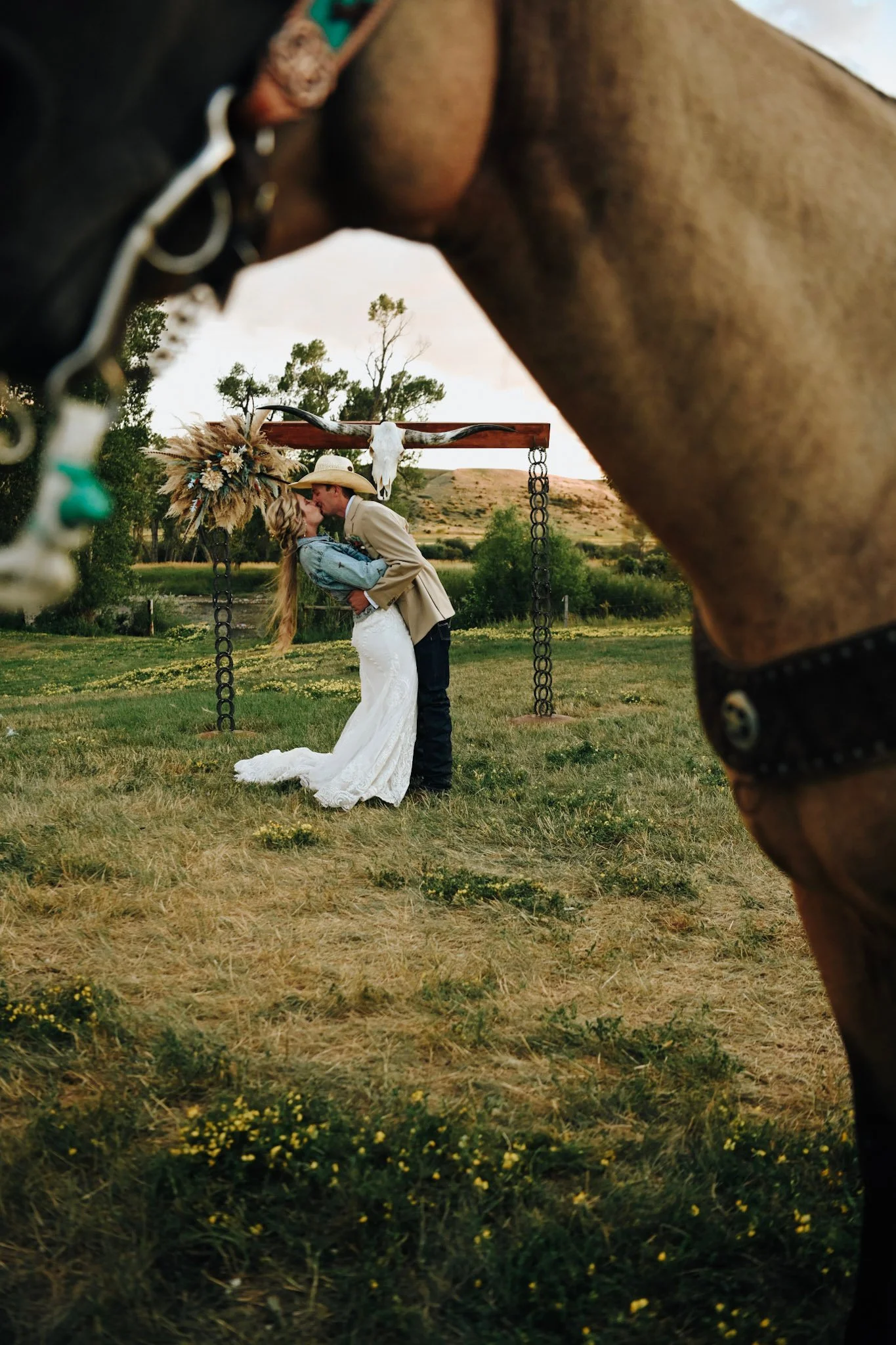 A couple wedding photo taken outdoors during sunset, with a horse's head in the foreground, framing the couple in the background. The couple is kissing, with the bride in a white dress and the groom in western attire, surrounded by a rustic setting.