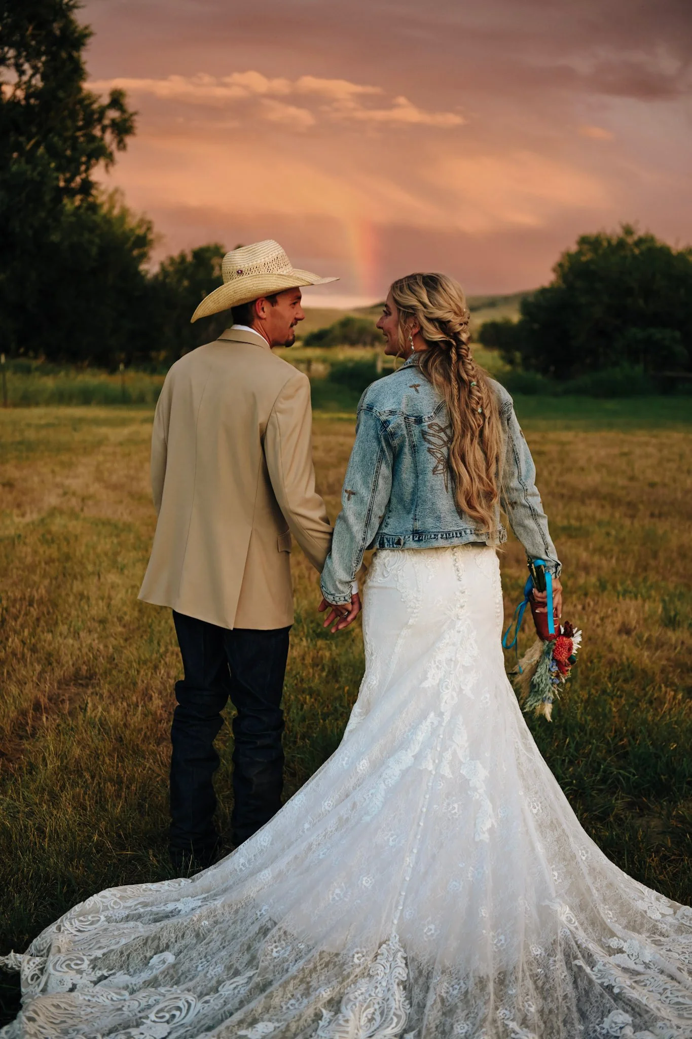 A couple holding hands in a field during sunset, with a rainbow in the sky, the woman is wearing a white lace wedding dress and the man is in a beige jacket and cowboy hat.