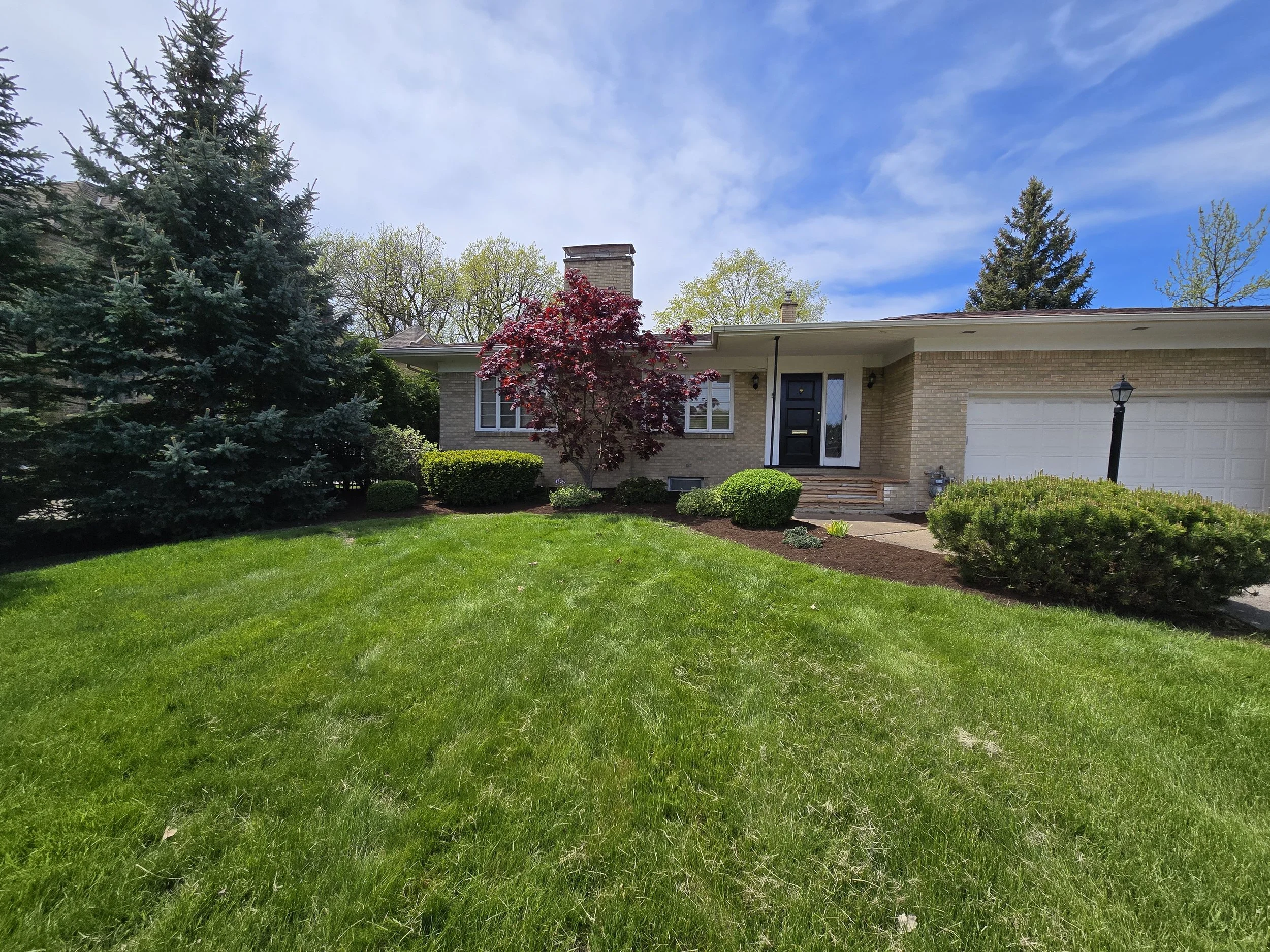 Front view of a house with a well-maintained green lawn, several trees including a large pine to the left, a red-leafed tree near the front porch, and a clear blue sky with some clouds.