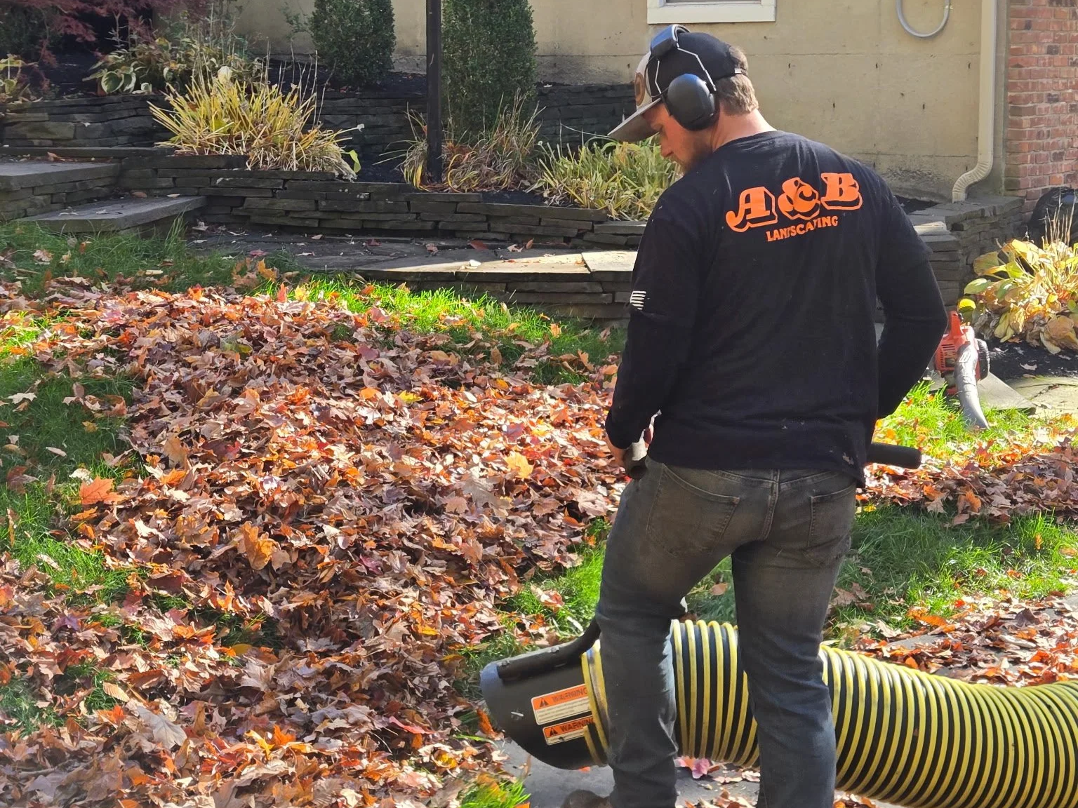 A landscaper wearing ear protection and a shirt with 'A&B Landscaping' works in a yard, blowing fallen autumn leaves into a pile with a leaf blower.