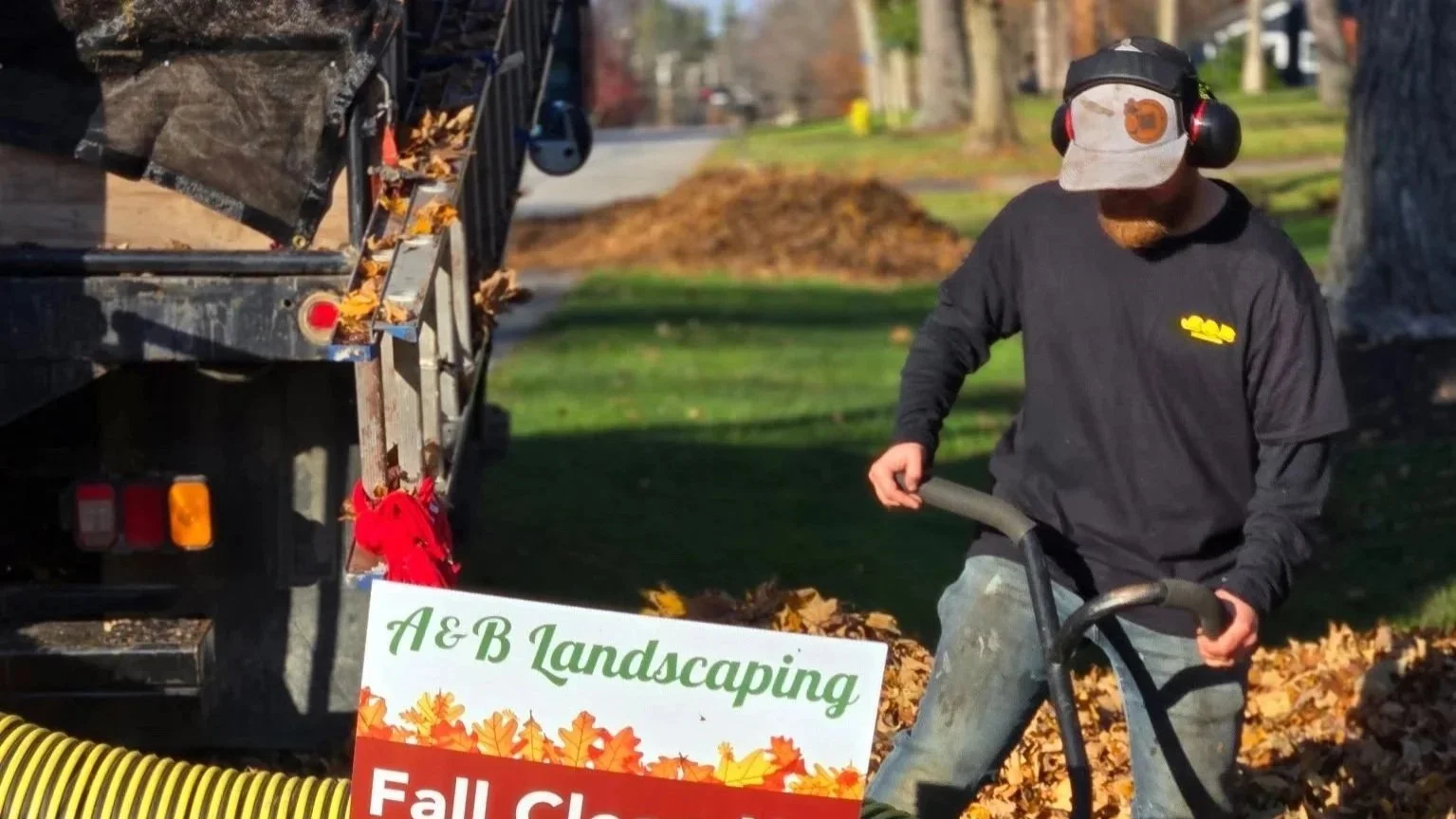 Lawn care worker wearing a black long-sleeve shirt, cap, and ear protection, using a leaf blower next to a truck with leaves, yard, and sidewalk in background.