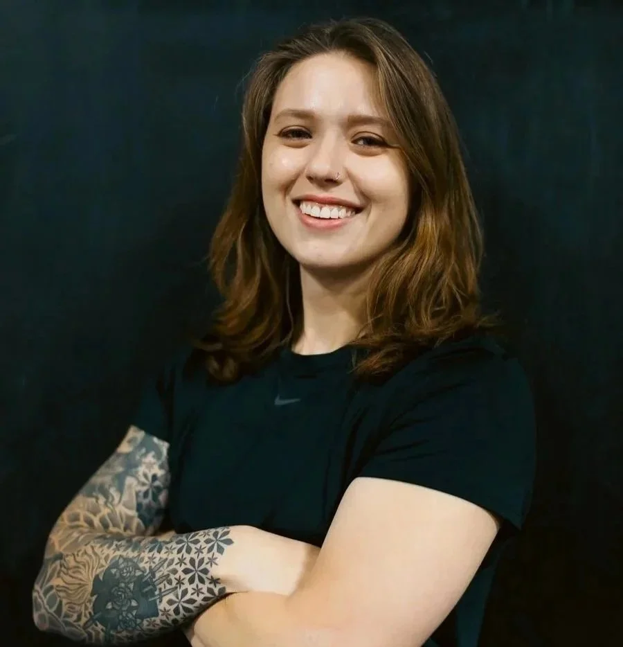 A smiling woman with shoulder-length brown hair, wearing a black short-sleeve athletic shirt, standing with arms crossed against a dark background. She has a tattoo sleeve on her left arm and a nose piercing.