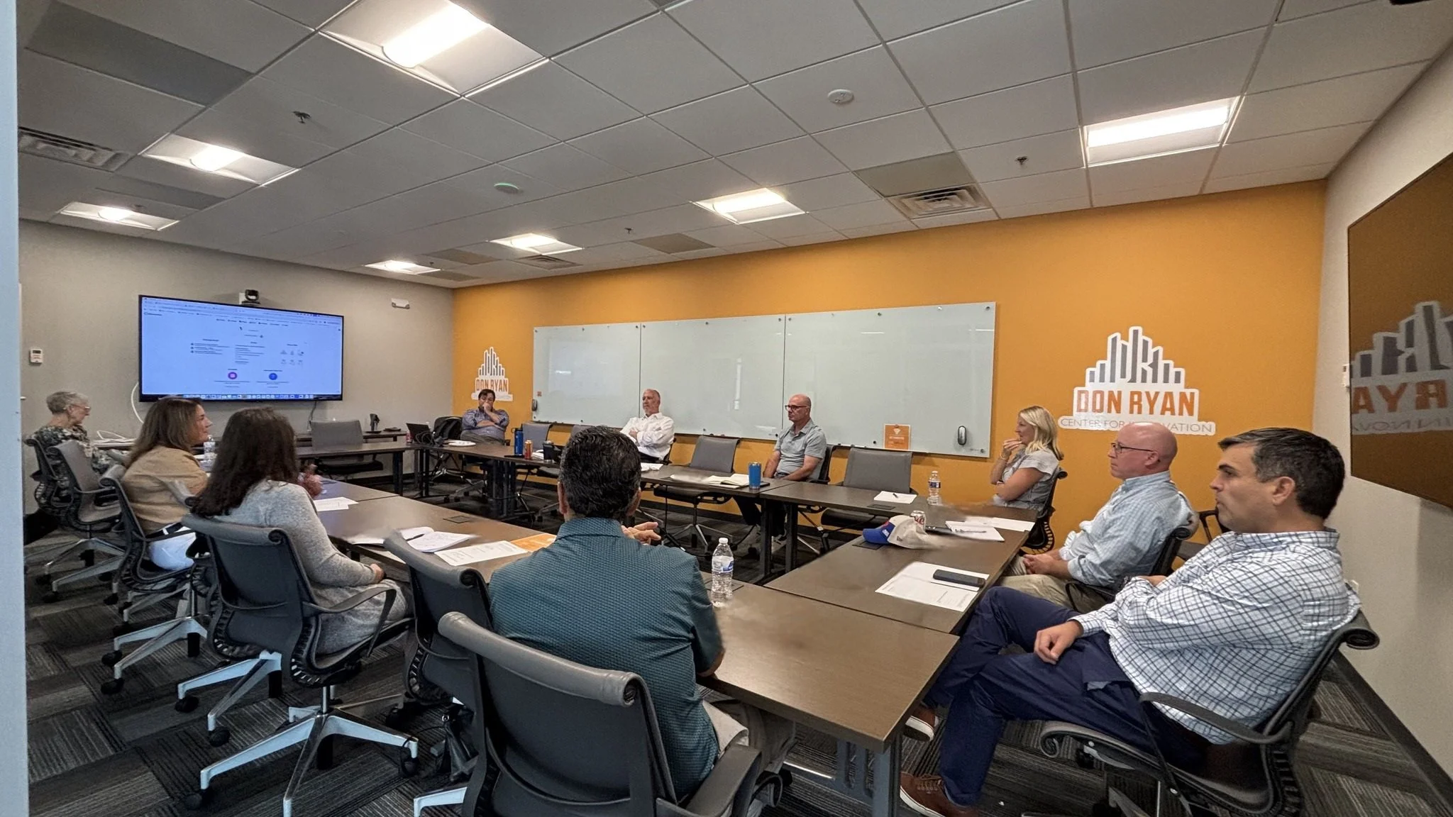 A group of people seated around a conference table in a meeting room with a whiteboard and a large screen displaying a presentation. There is a sign on the wall that says 'DON RYAN CENTER FOR INFORMATION'.