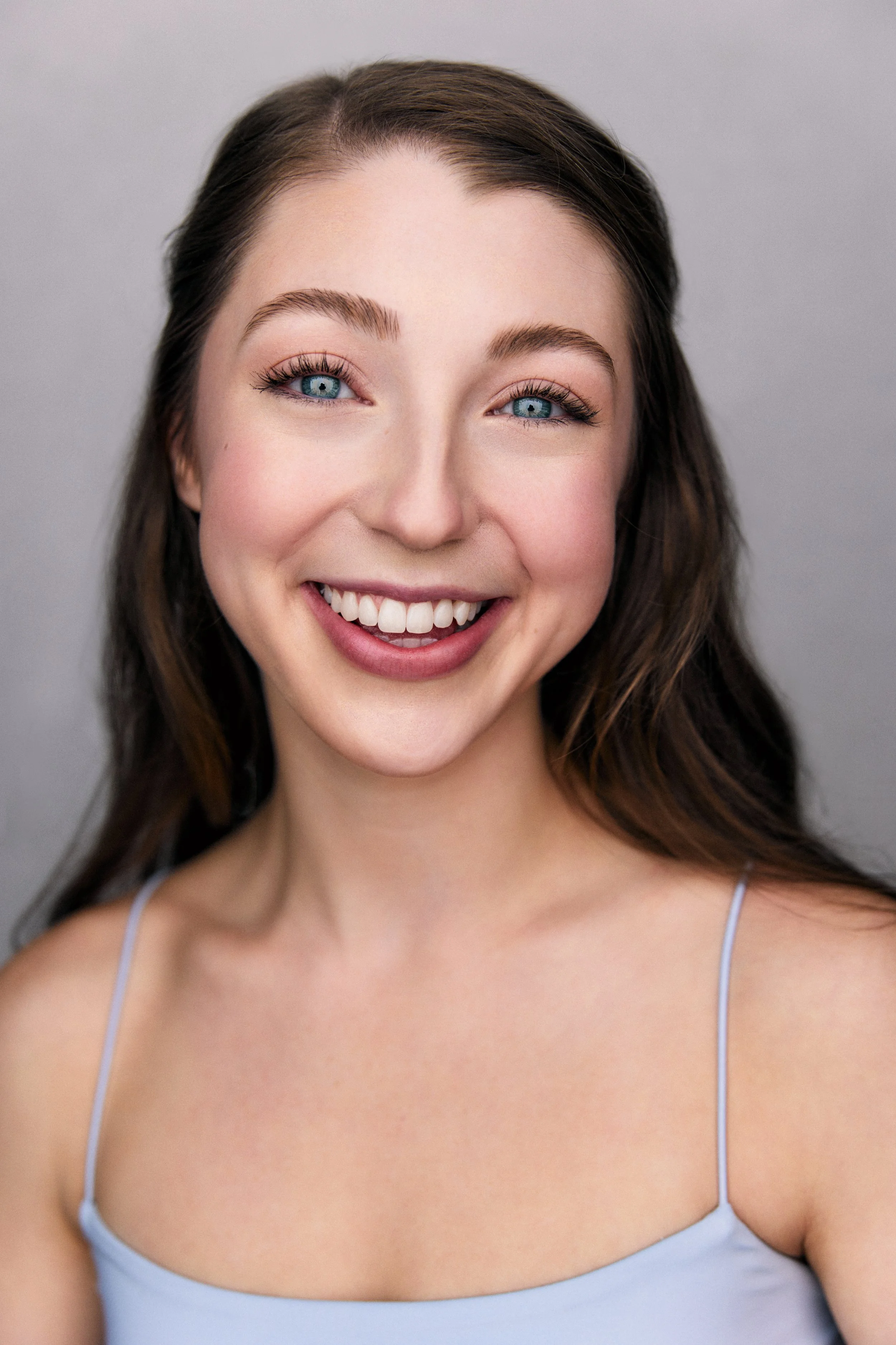 Close-up portrait of a smiling young woman with blue eyes, light makeup, and long brown hair, wearing a light blue spaghetti strap top, against a plain gray background.
