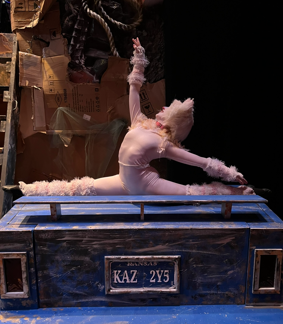 A performer in a white bodysuit with fluffy pink accessories on arms and legs, and a white wig, is doing a split on a blue platform during a show.
