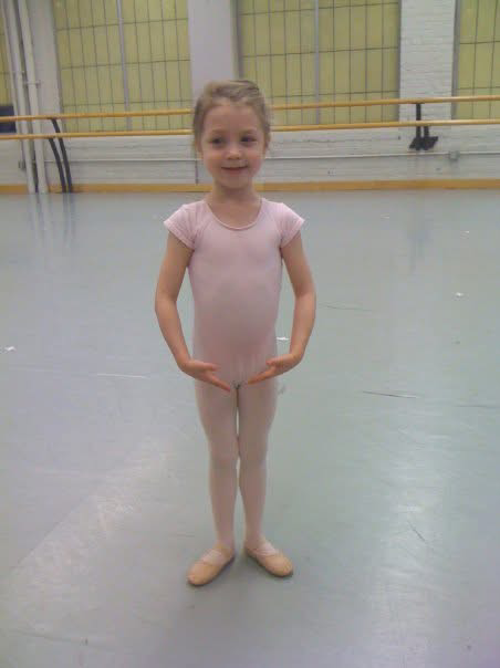 A young girl in a pink ballet outfit standing in a dance studio with ballet barres.