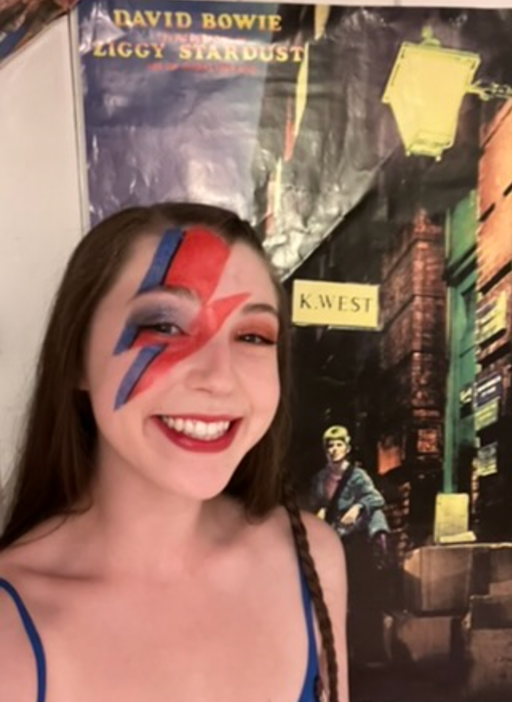 A young woman with a big smile, face painted with a red, blue, and white lightning bolt design, standing in front of a wall poster for David Bowie and Ziggy Stardust