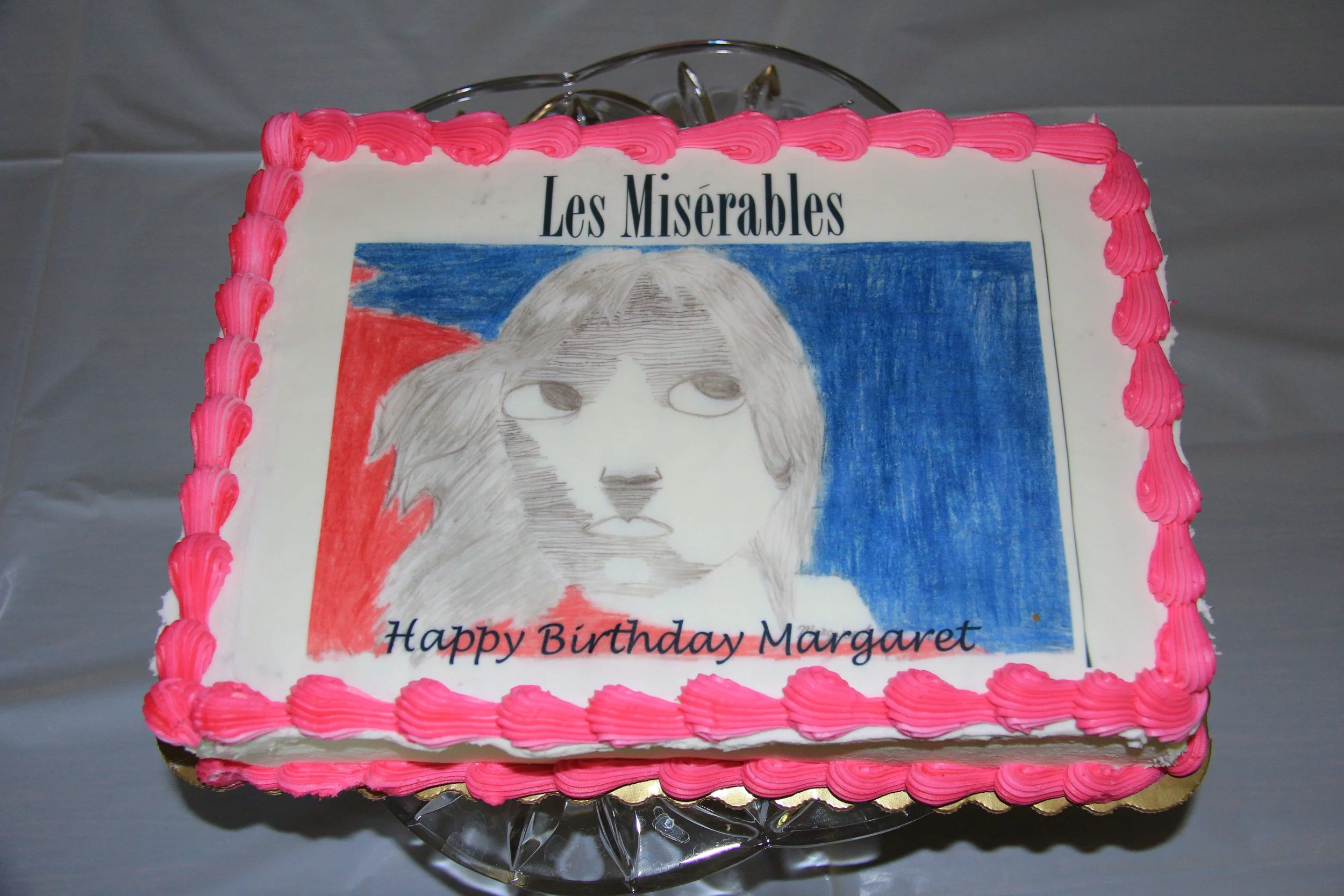 A birthday cake with an edible image of the musical Les Misérables, with a pink and white frosting border and the words 'Happy Birthday Margaret' written on it.