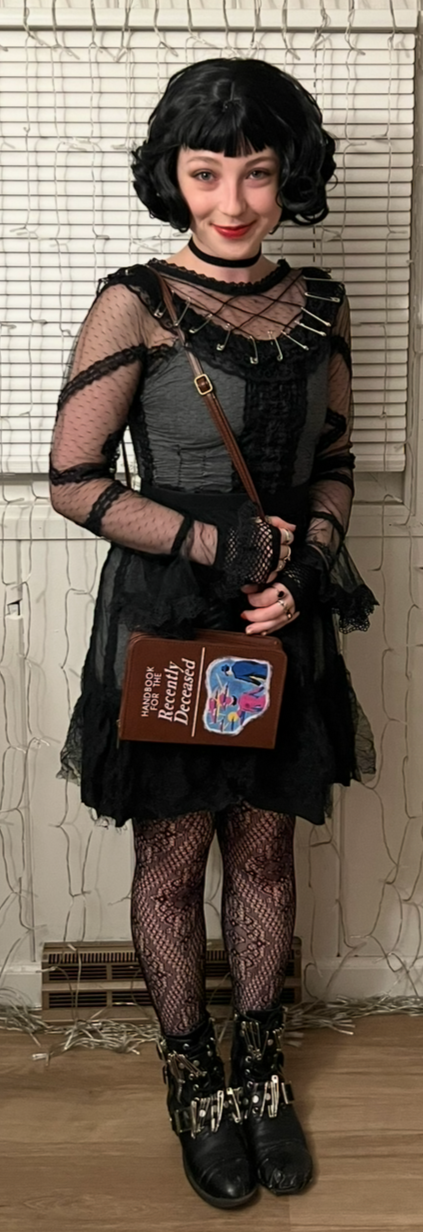 Woman dressed in a gothic punk style outfit with black curly hair, sheer black top, black skirt, fishnet tights, combat boots with buckles, and a choker, standing in front of a window with blinds.