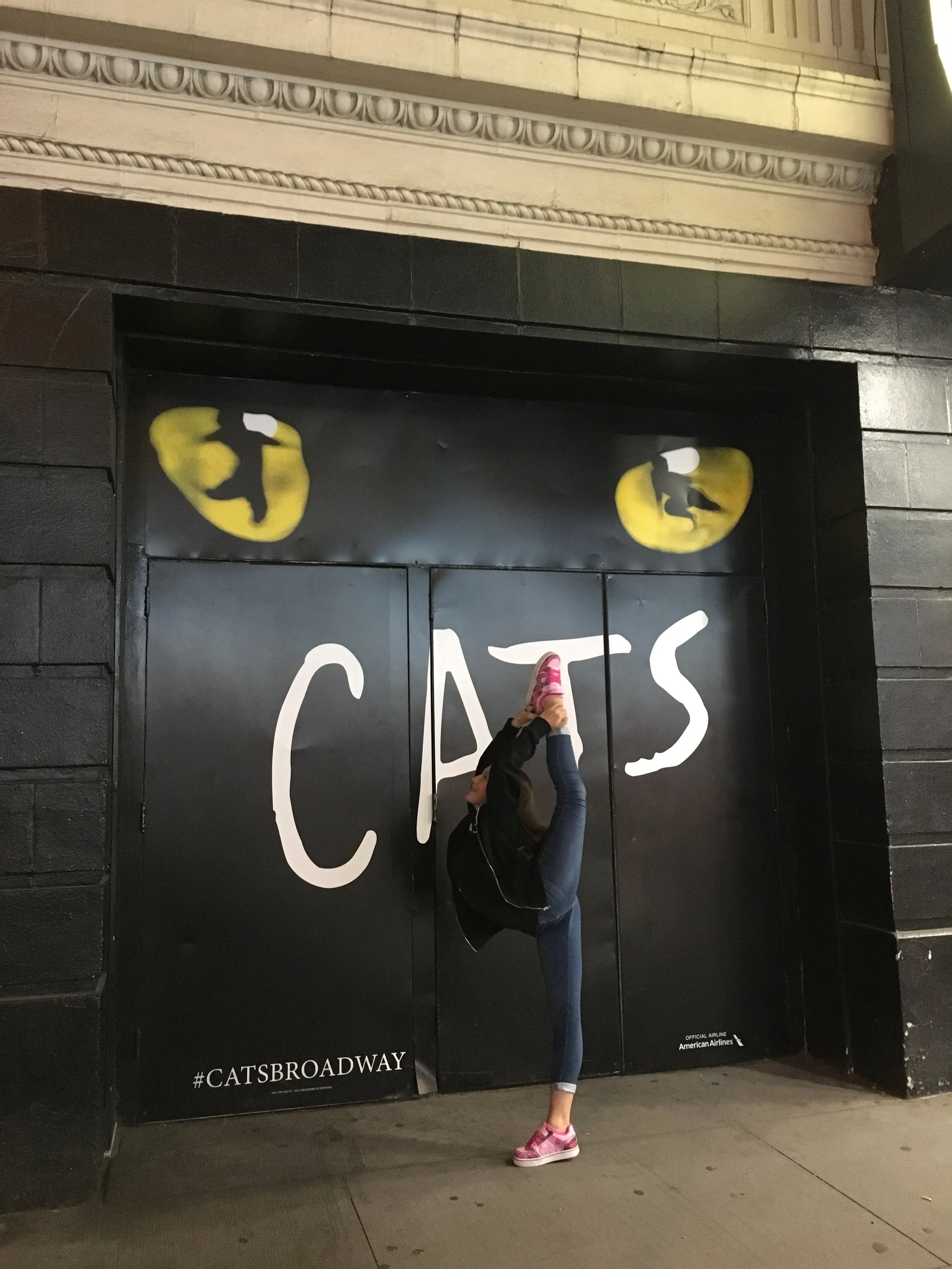 A young woman with a high leg stretch against a black wall with a painted 'CATS' sign and yellow cat eyes on top, performing a high kick.