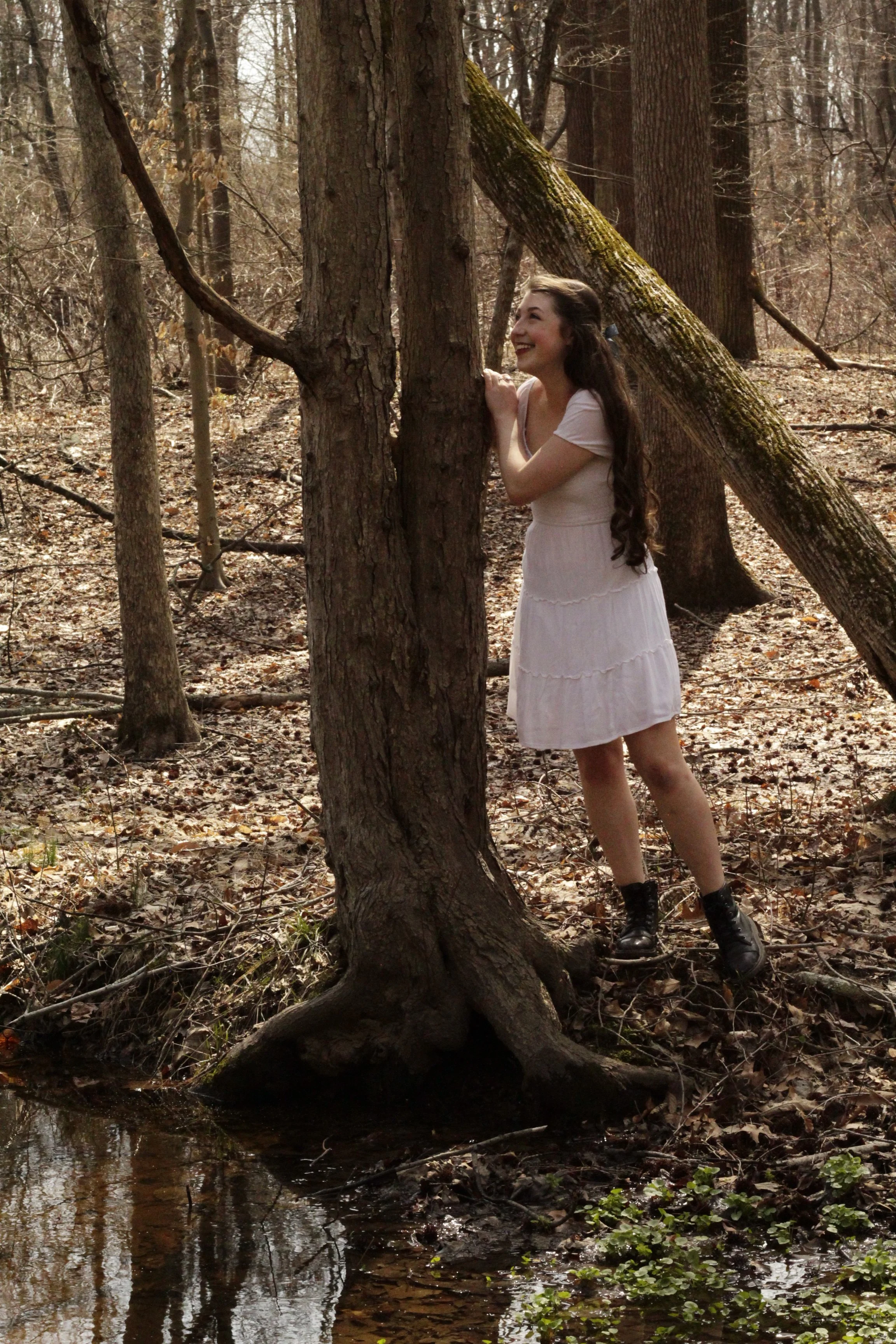 Young woman in a white dress and black boots hugging a tree in a forest during fall, with a small stream in the foreground.