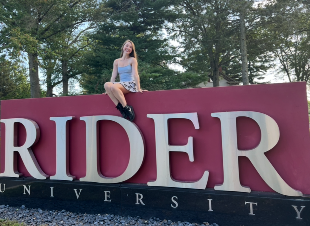 A young woman sitting on top of a sign that reads 'RIDER UNIVERSITY,' with a background of trees and a partly cloudy sky.