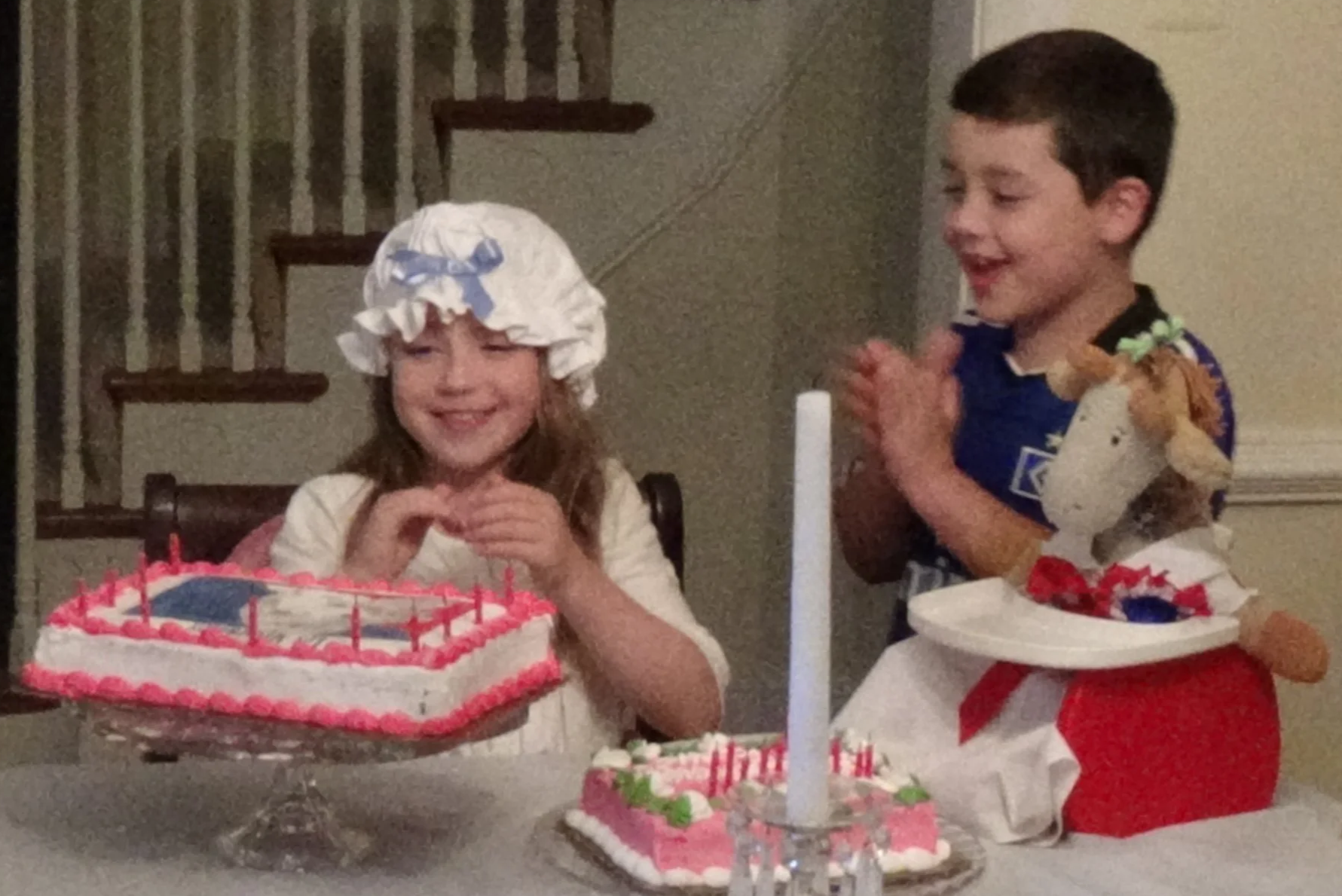 Two children celebrating a birthday with decorated cakes, one girl wearing a white bonnet with a blue bow, and a boy wearing a dark shirt. The girl is smiling while the boy claps. There are candles on the cakes and a lit candle in a candle holder on the table.
