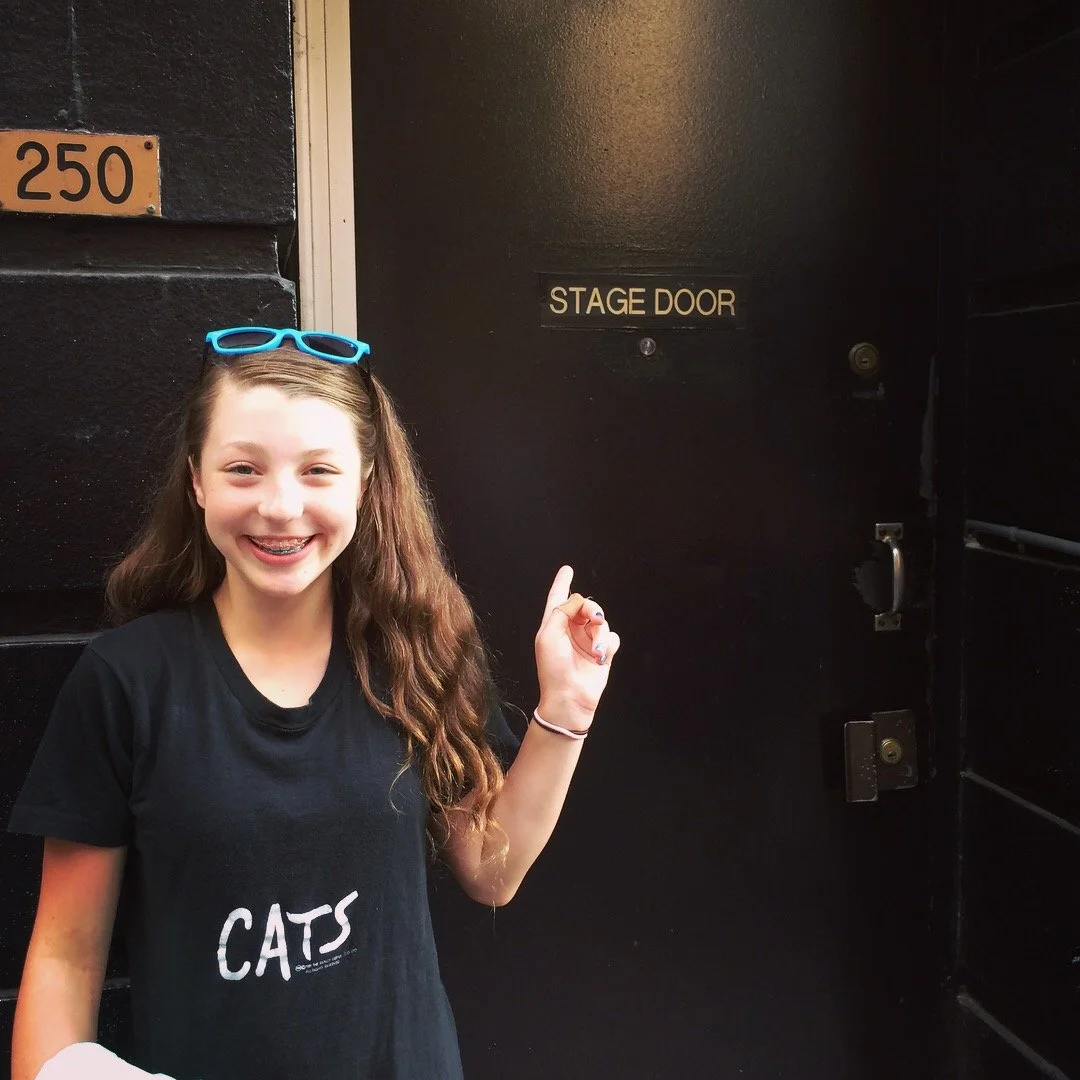 A smiling girl with long hair, wearing glasses on her head and a black t-shirt with 'CATS' written on it, standing in front of a black stage door.