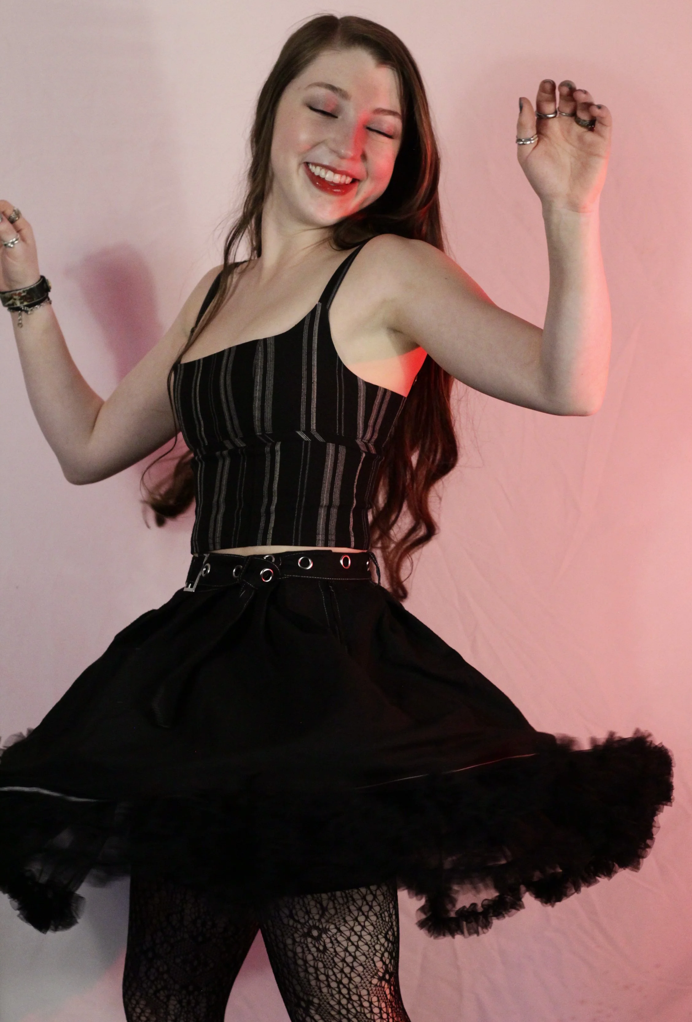 A young woman with long brown hair is smiling with her eyes closed, wearing a black striped top, a black tutu skirt, fishnet stockings, and multiple rings and bracelets.