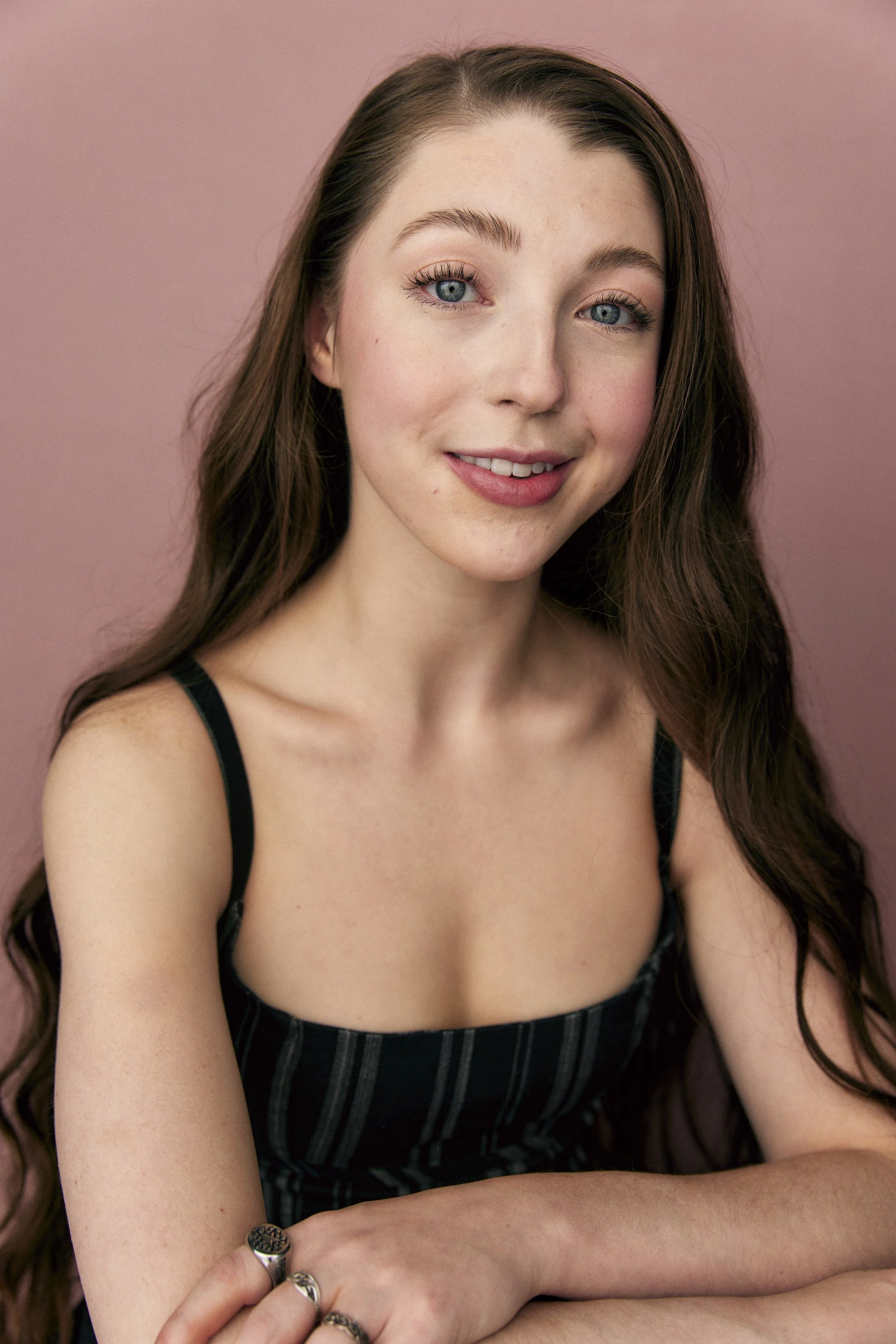 A young woman with long brown hair and blue eyes smiling, wearing a black and gray striped sleeveless top, against a pink background.