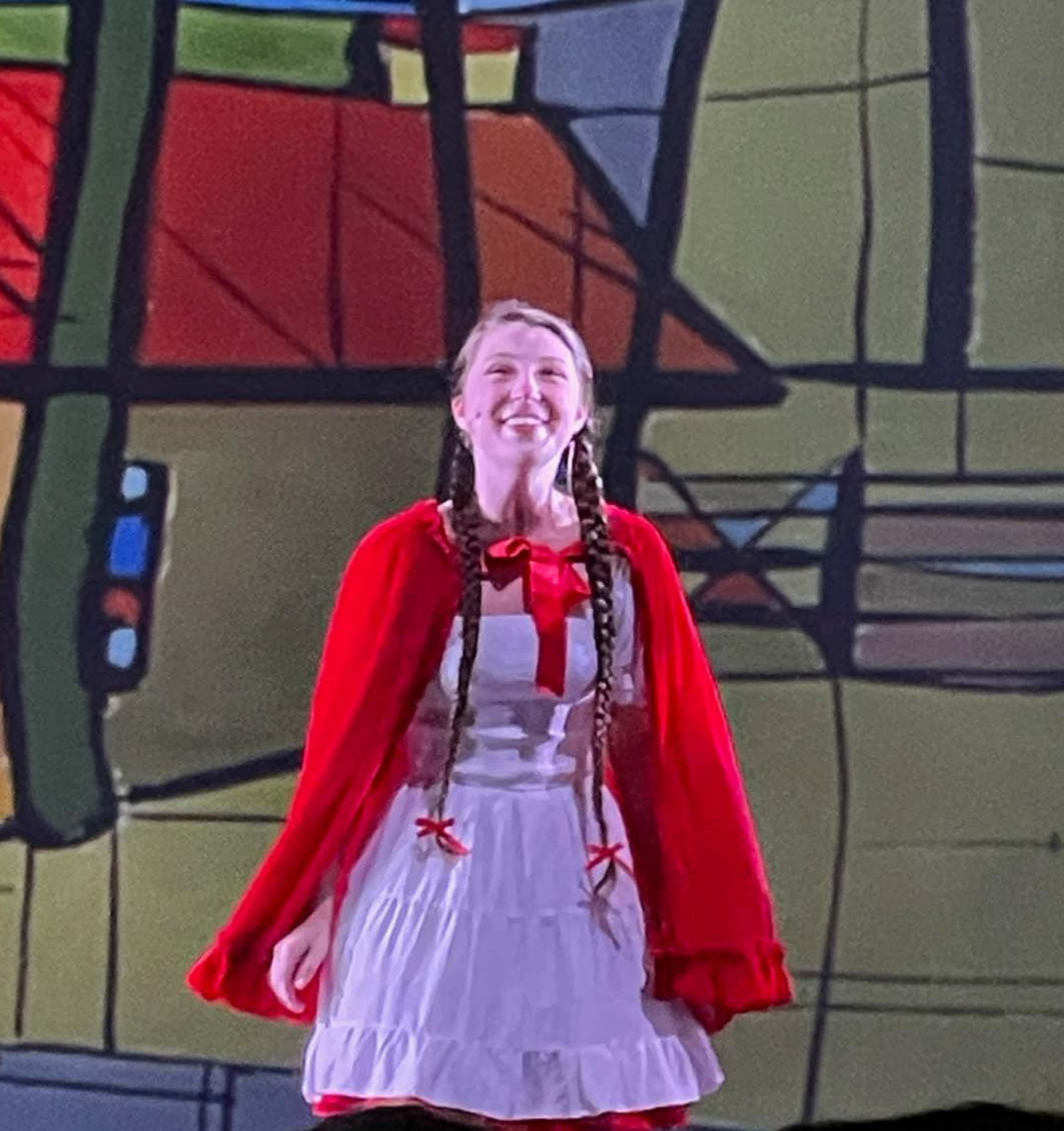 A girl in a white dress with red accents and a red cape, smiling on stage with a colorful abstract background.