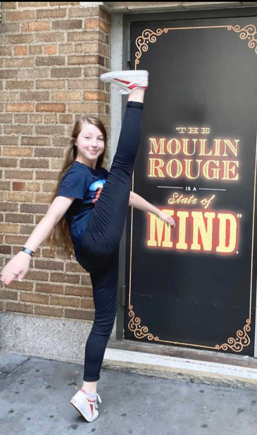 Young girl doing a high kick in front of a sign that reads 'The Moulin Rouge is a State of Mind'.