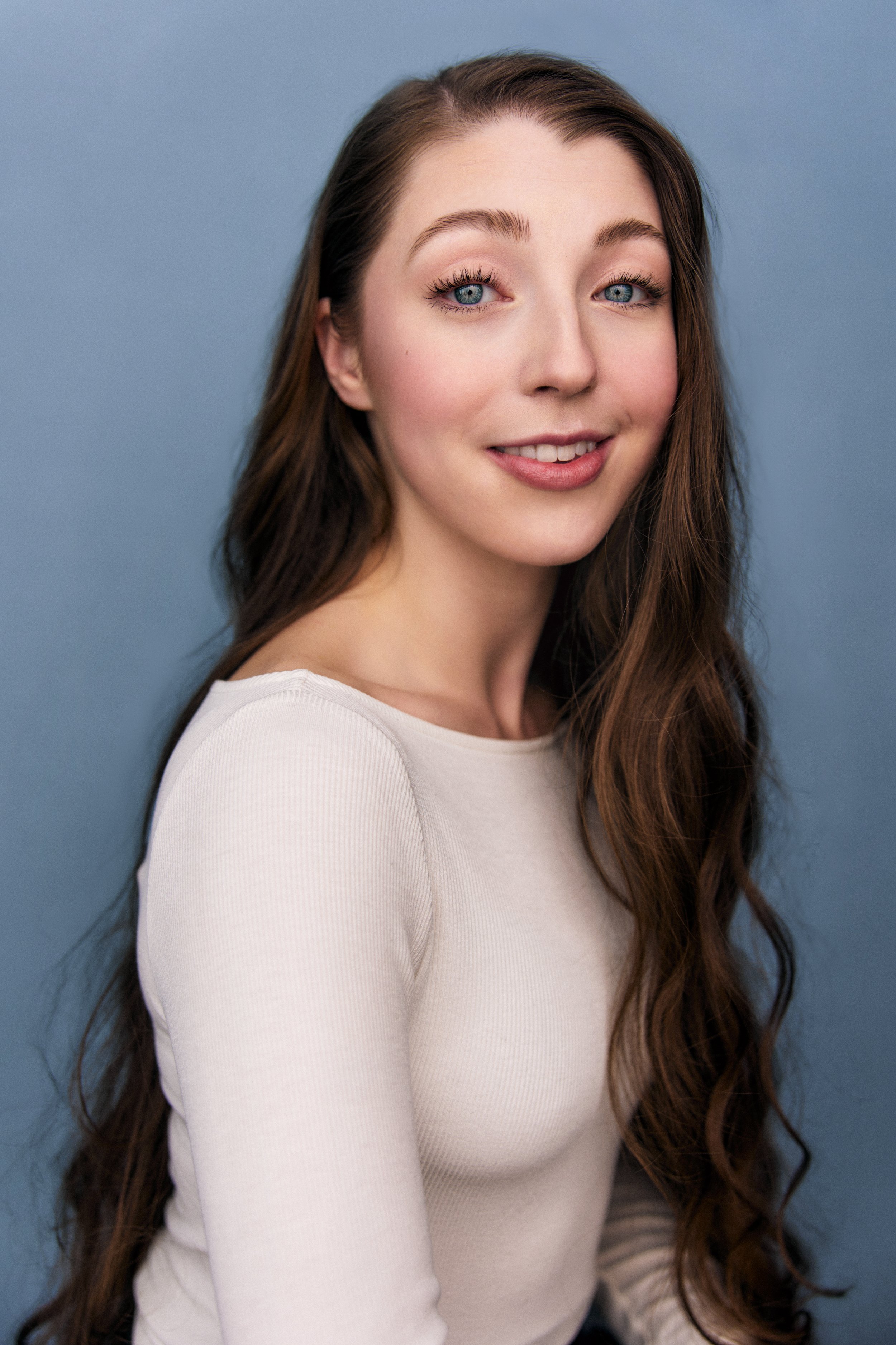 A young woman with long, wavy brown hair and blue eyes smiling softly against a blue background.